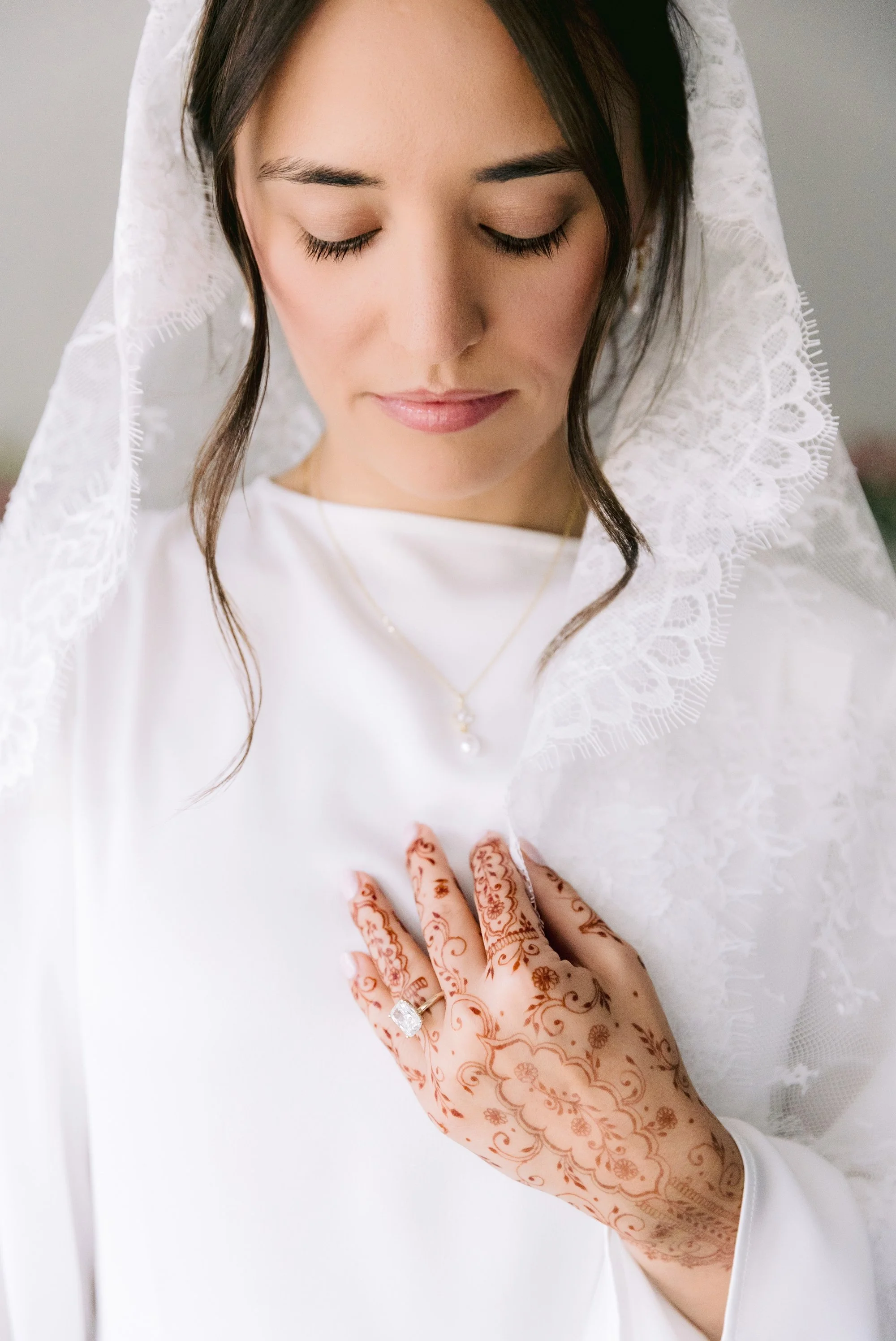 A woman with closed eyes and a white lace veil looking downward with her hand on a white garment, decorated with henna designs and a ring.