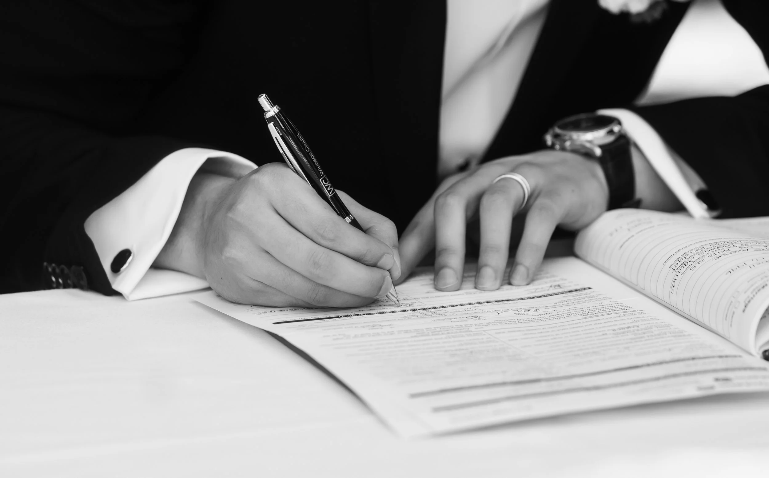 A person dressed in formal attire is signing a document with a pen, with an open book nearby, on a desk.