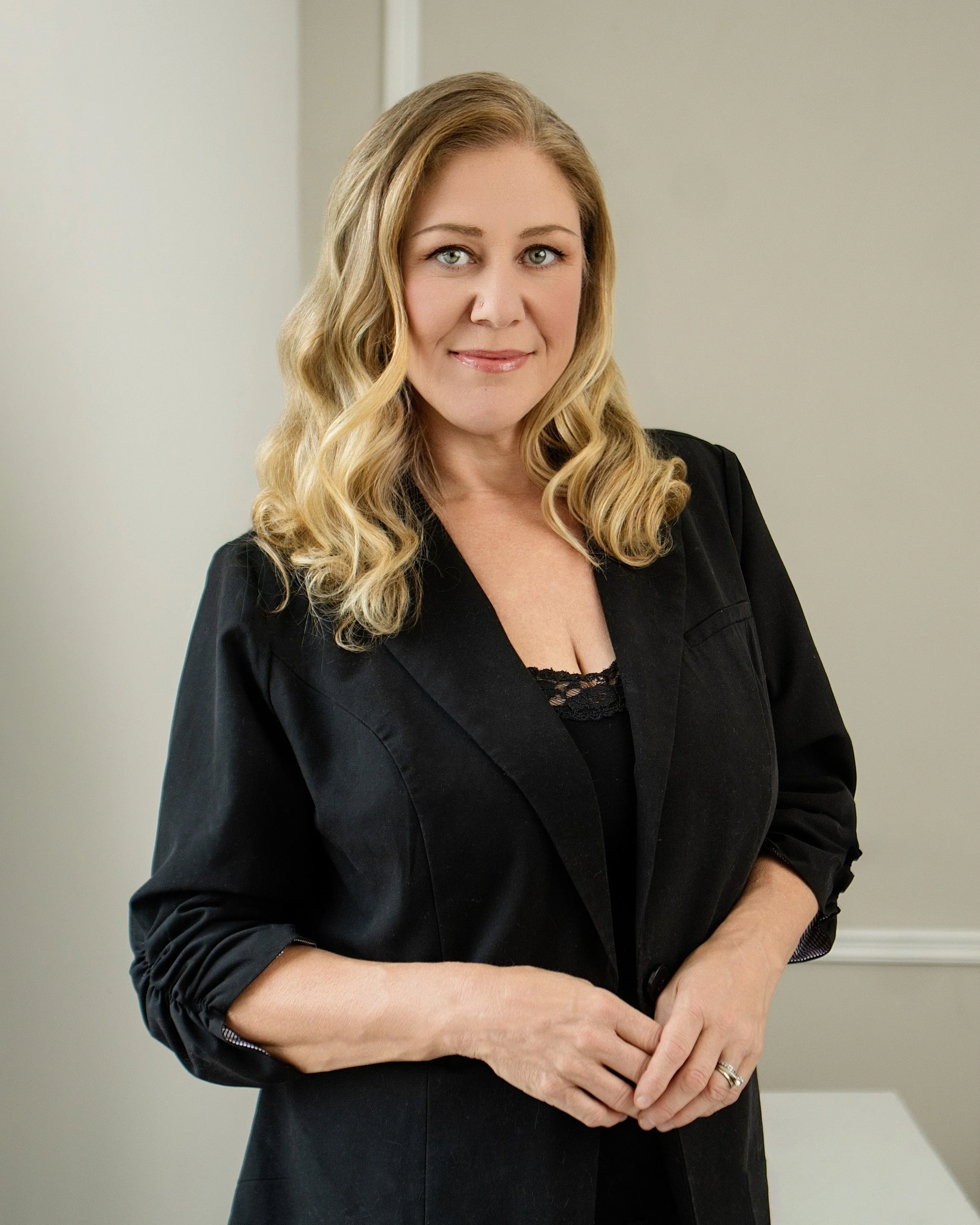 A woman with long blonde hair, light skin, and gray eyes dressed in a black blazer, standing against a plain neutral wall, arm crossed, smiling subtly.