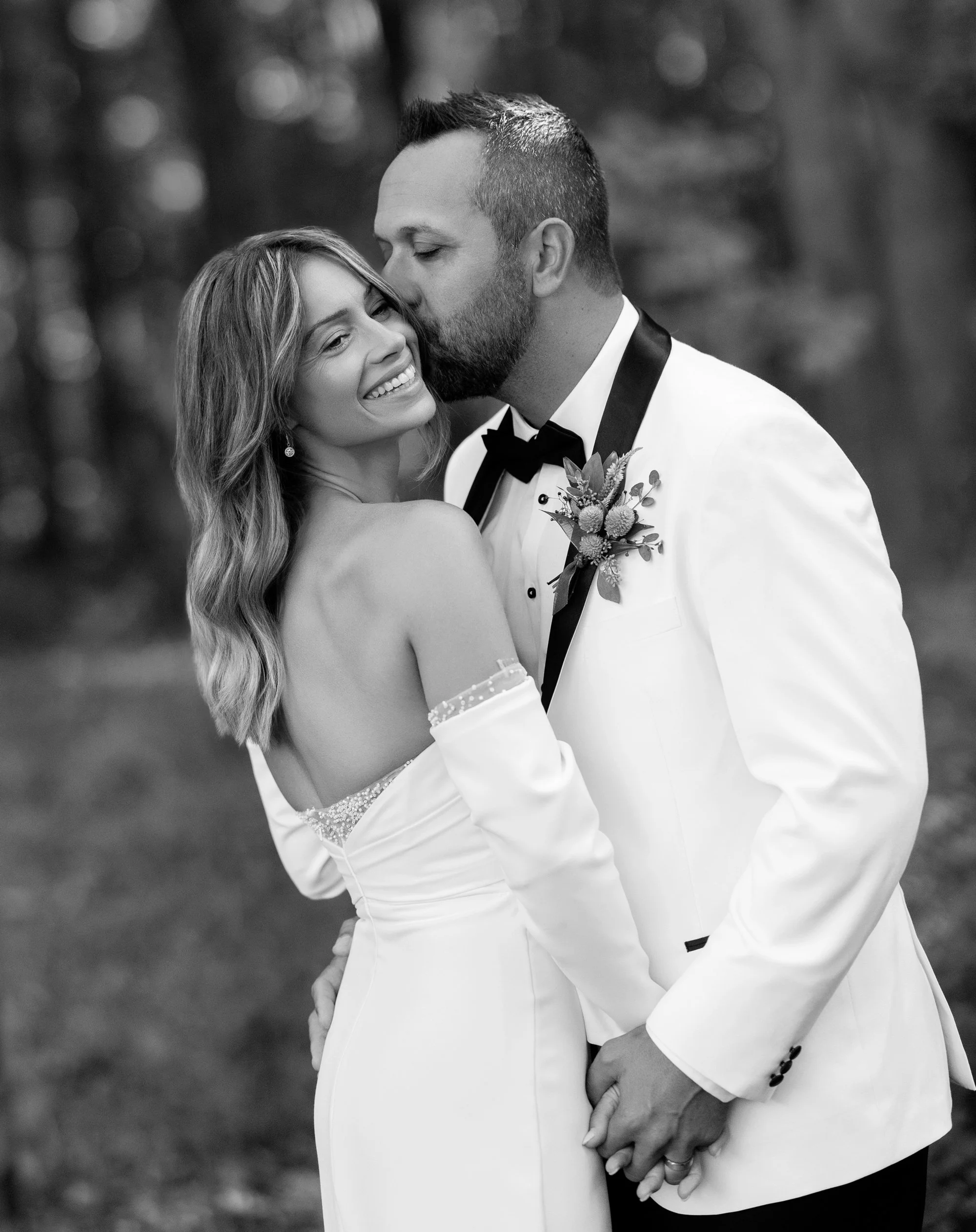 Black and white photo of a bride and groom embracing outdoors, with the groom kissing the bride's temple. The bride is smiling with her eyes closed, showing her wedding dress and wavy hair. The groom is wearing a white tuxedo jacket with black accents and boutonniere, and has neatly styled hair and beard.