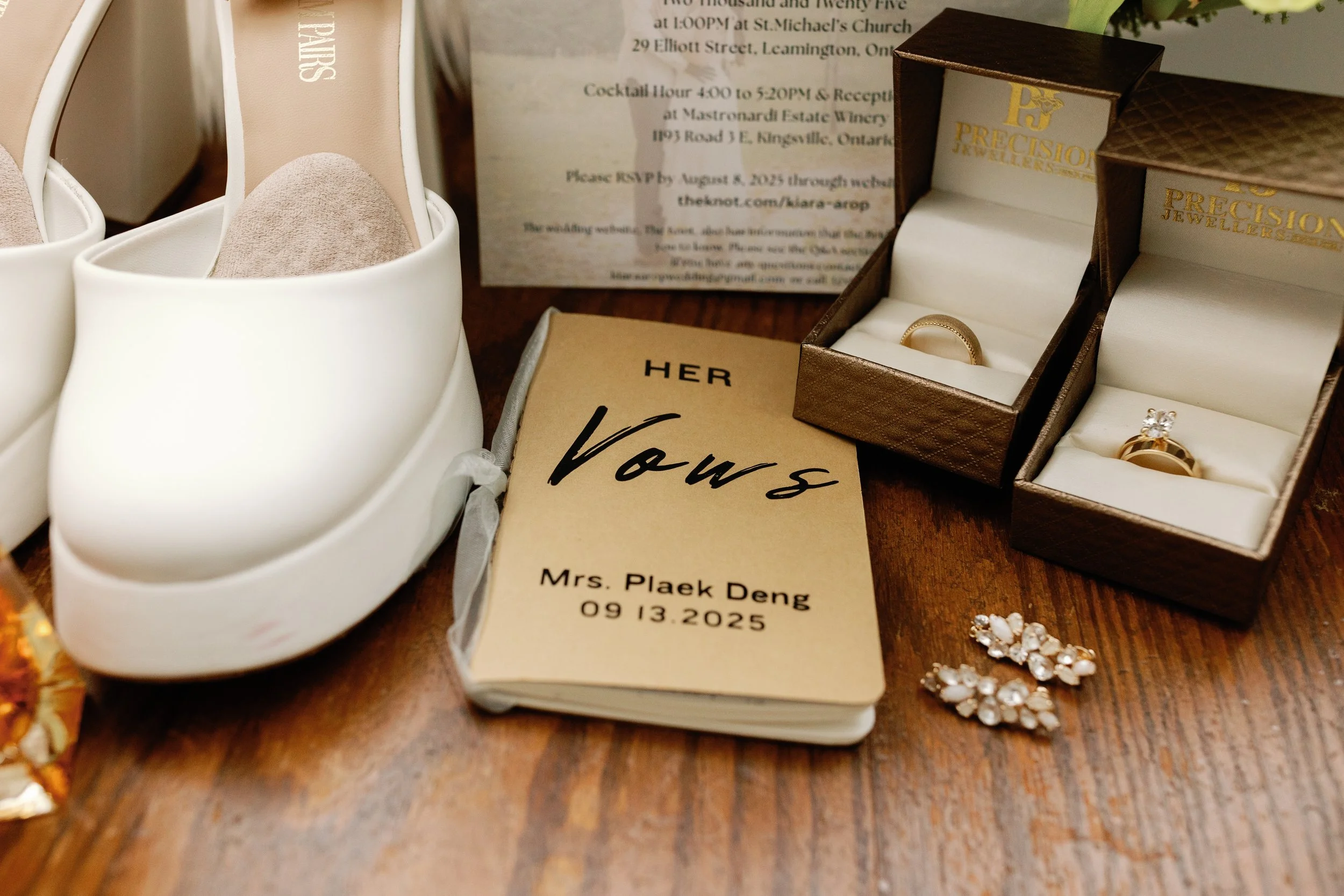A collection of wedding items including white shoes, a wedding ring in a brown box, pearl jewelry, a wedding invitation card, and an RSVP card, all displayed on a wooden surface.