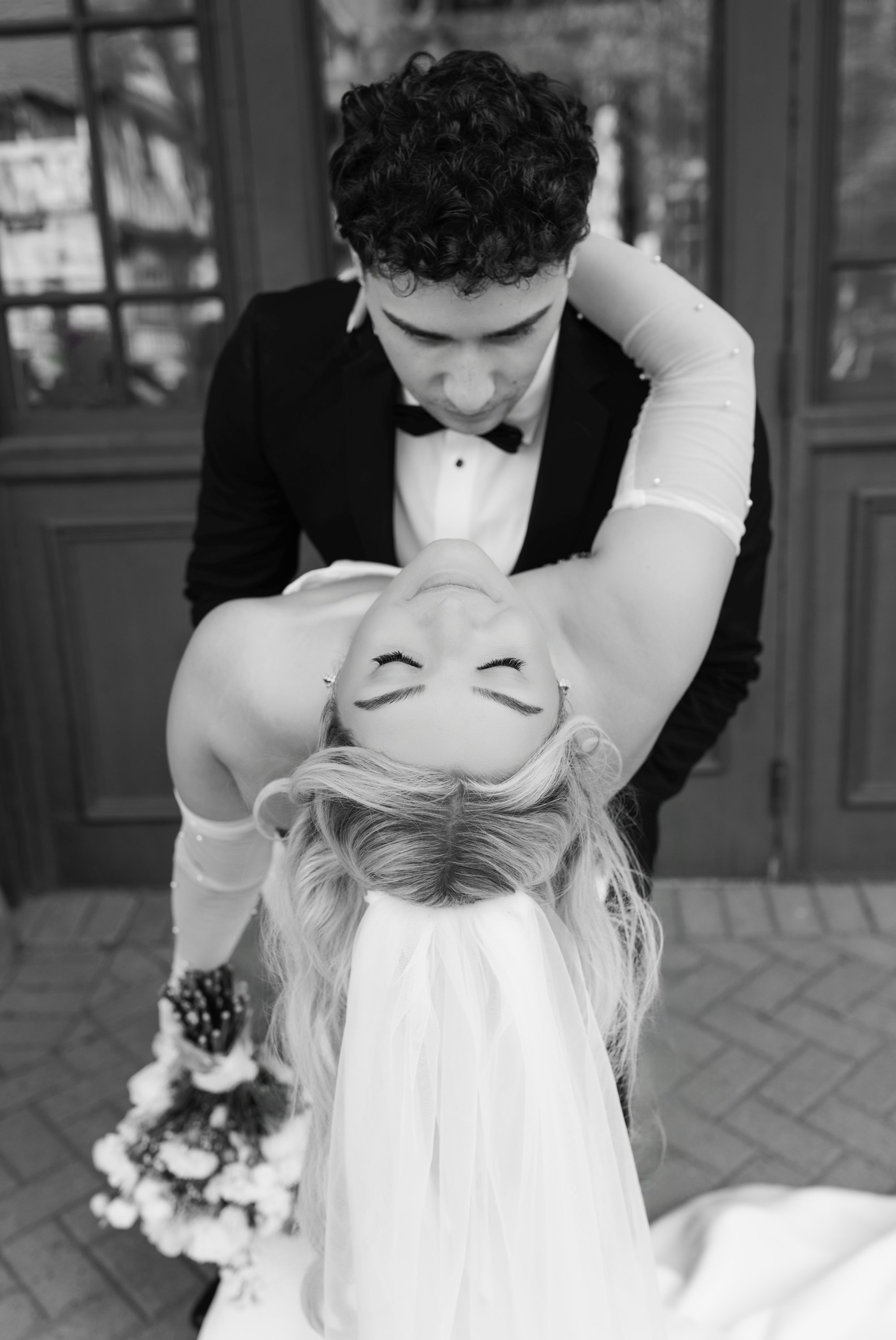 A man in a tuxedo and bow tie dances with a woman in a wedding dress, with a bouquet of flowers in the background.