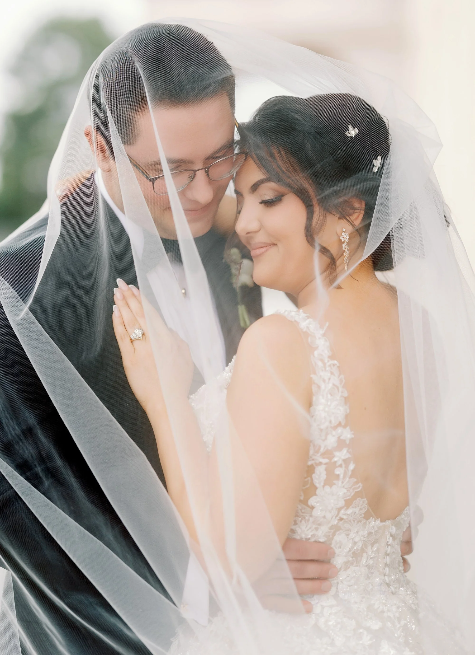 A bride and groom sharing a close, intimate moment under a veil, with the bride wearing a lace wedding dress and statement earrings, and the groom in a black tuxedo with glasses.