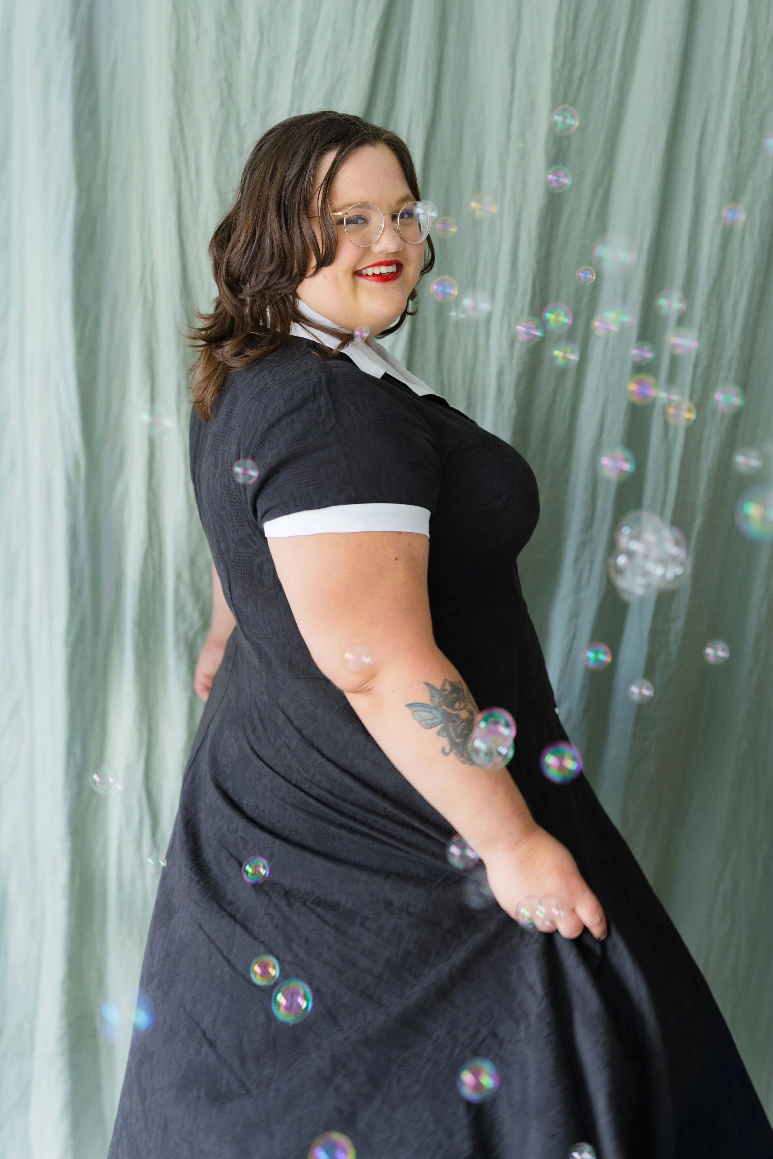 A woman with shoulder-length brown hair, glasses, red lipstick, and a tattoo on her arm, dressed in a black dress with a white collar, standing in front of a green curtain with soap bubbles floating around her.