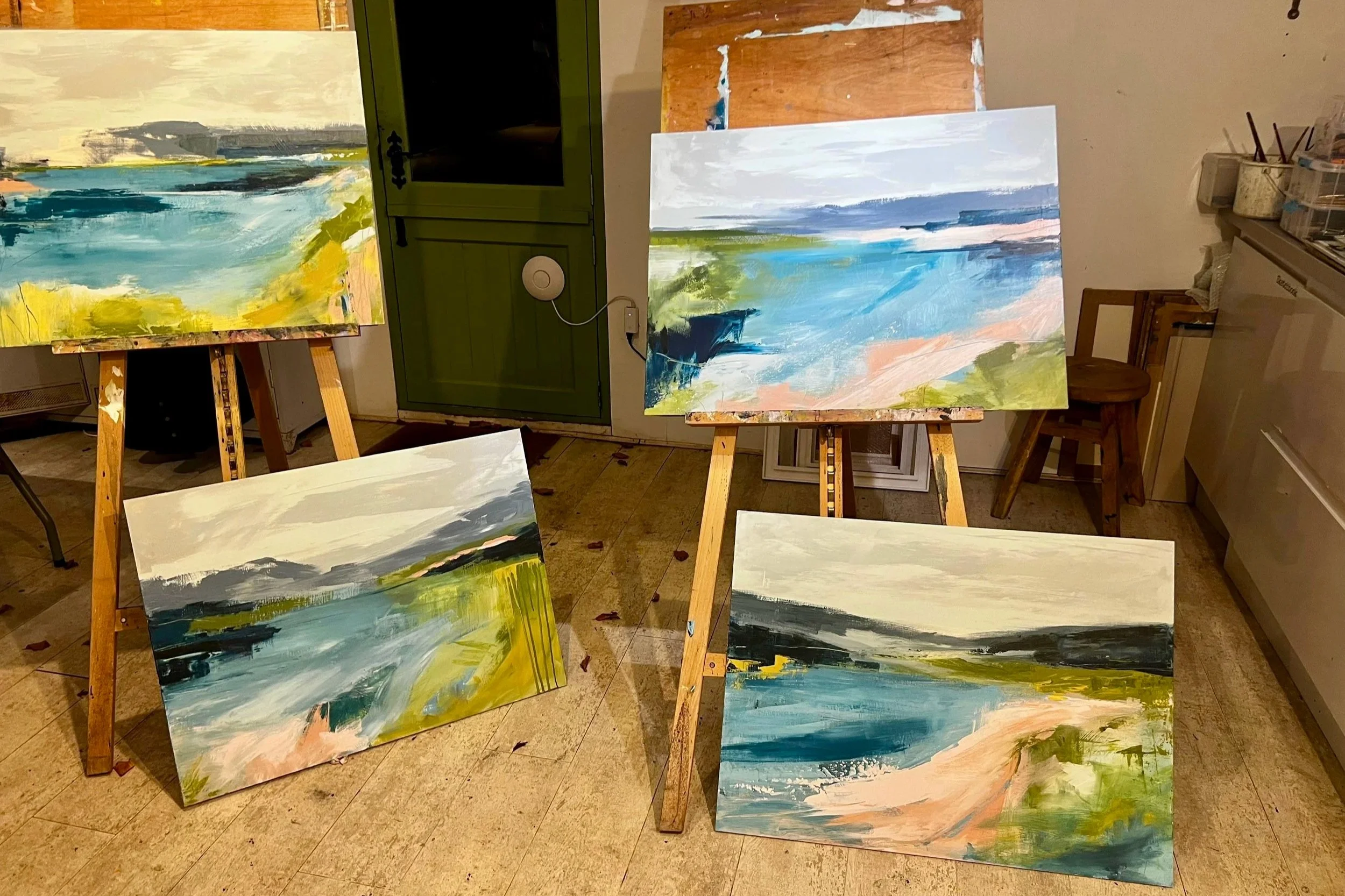 Multiple abstract landscape paintings on easels in an art studio, with landscape elements like sky, water, and greenery depicted in bold brushstrokes.