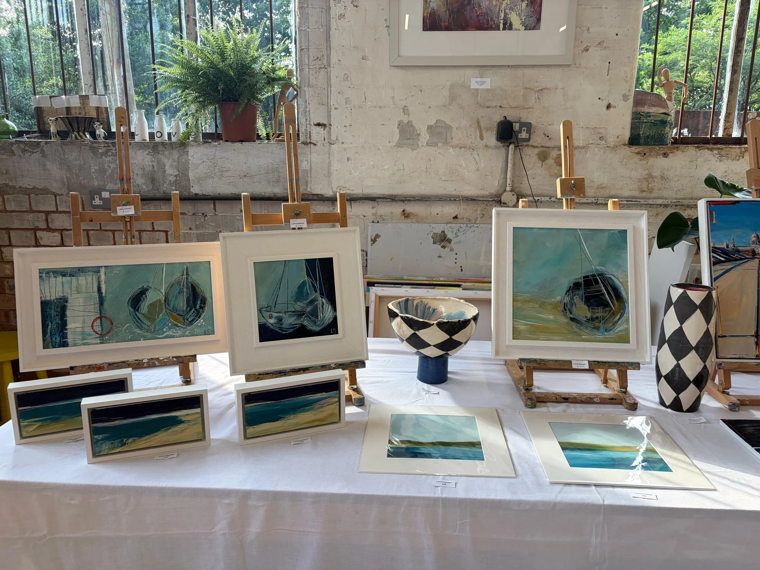 Artwork display at an art gallery featuring abstract paintings of maritime scenes with the ocean and boats, arranged on tables and easels, with plants and windows in the background.