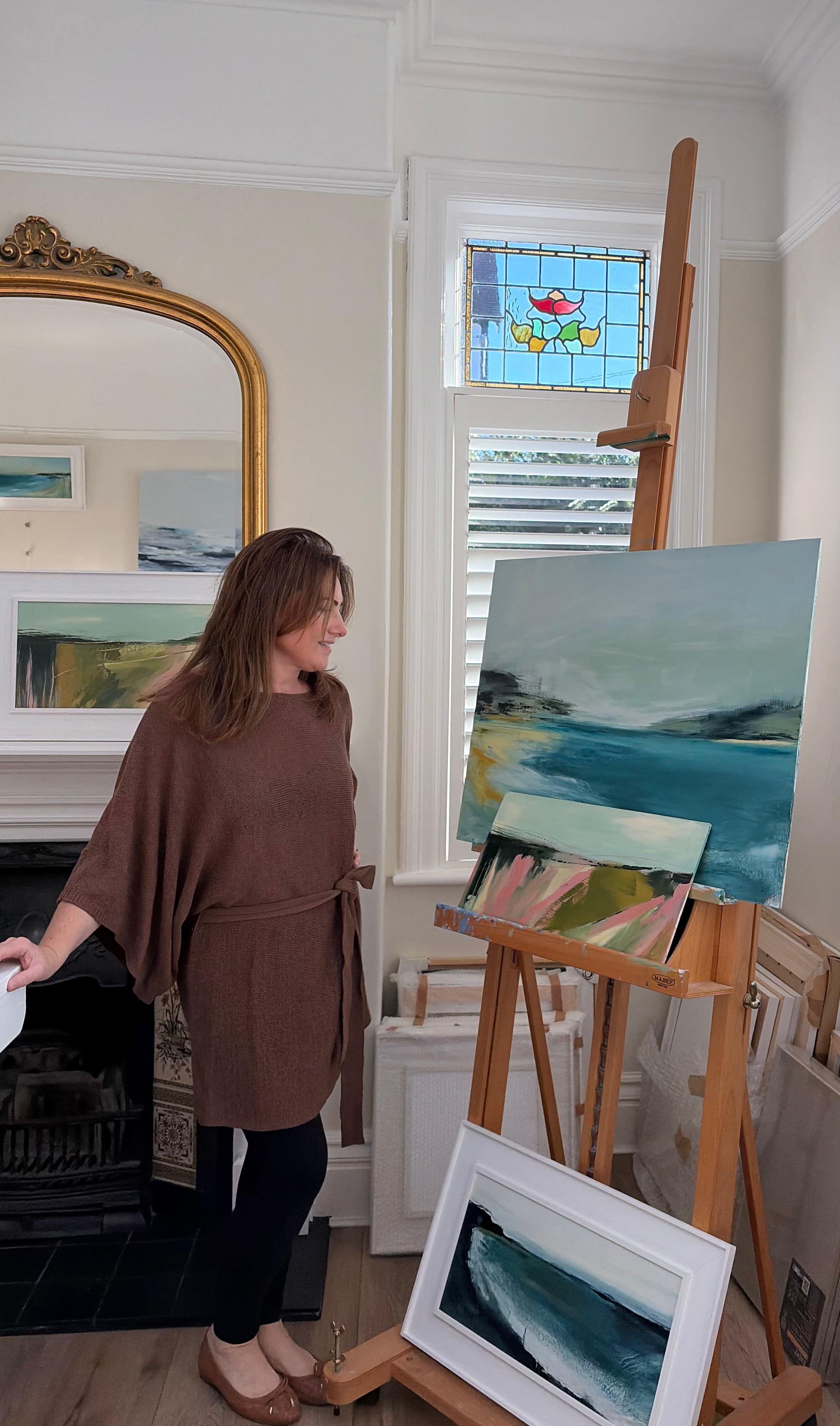 A woman in a brown dress and flats stands in front of a large landscape painting on a wooden easel, inside a room with white walls, a gold-framed mirror, a window with a stained glass design, and other framed artwork.