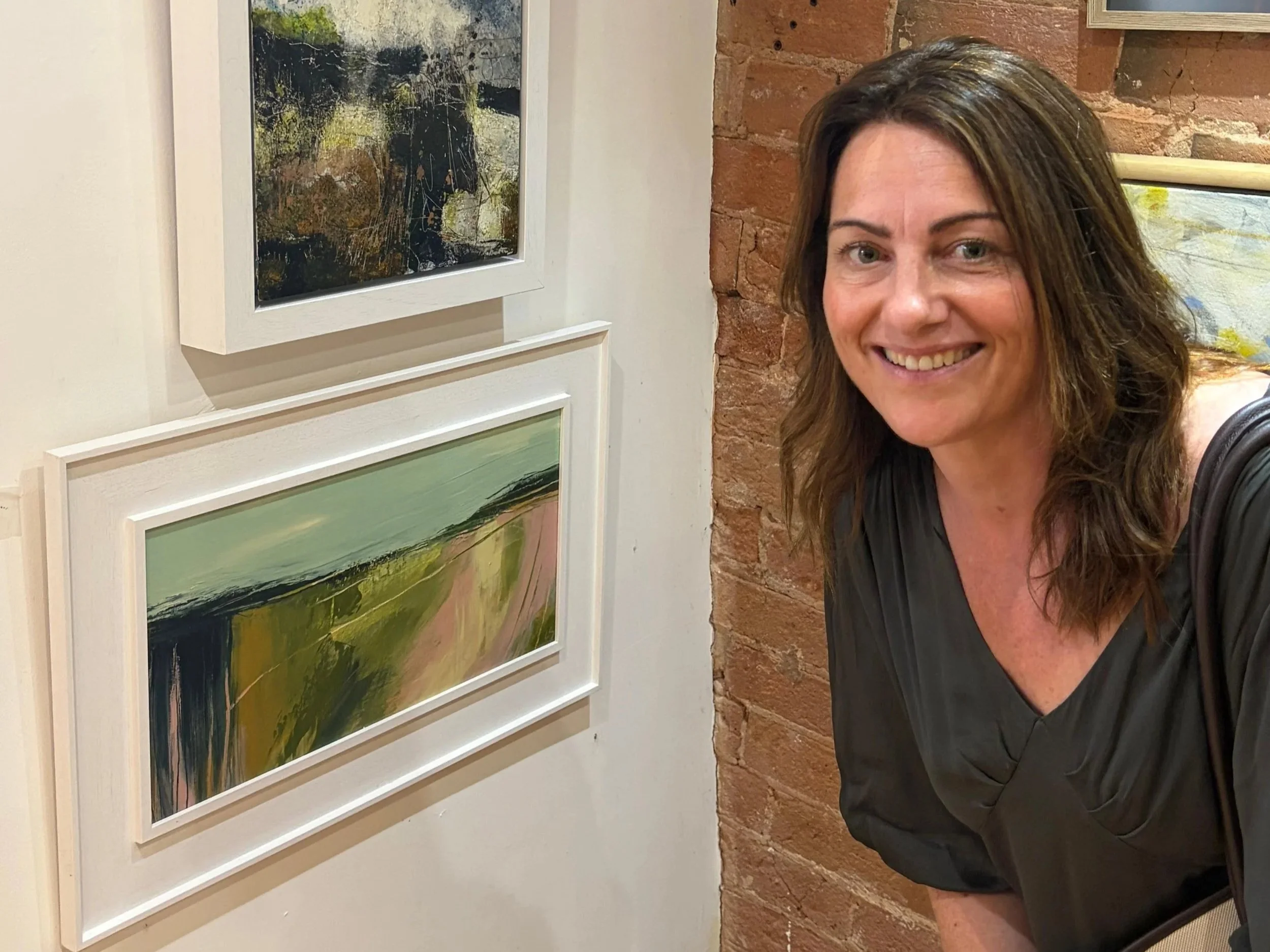 Lorna Stewart smiling, wearing a black t-shirt, standing next to a display of three abstract landscape paintings on a white wall, with a brick wall in the background.