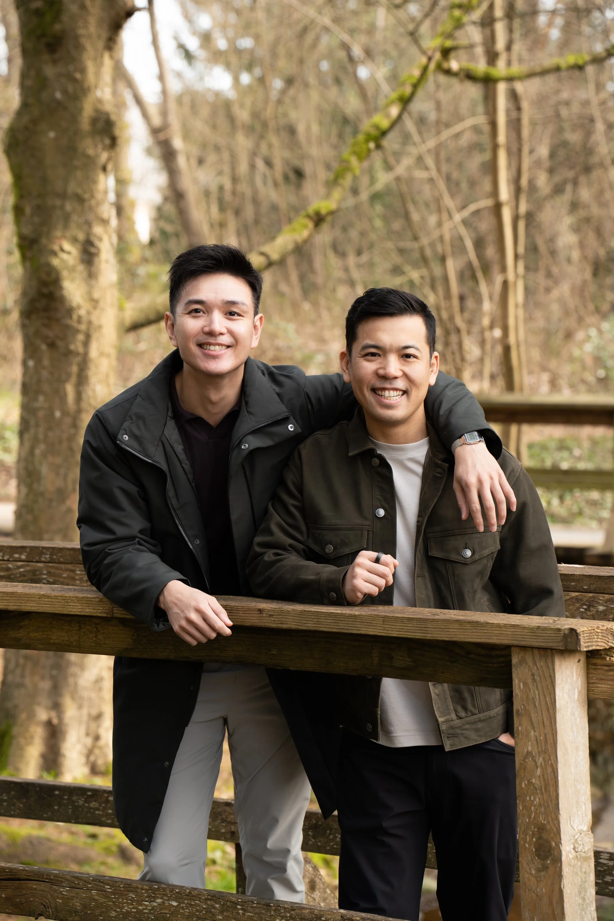 Two smiling men with dark hair in casual outdoor clothing standing together behind a wooden fence in a wooded area during daytime.