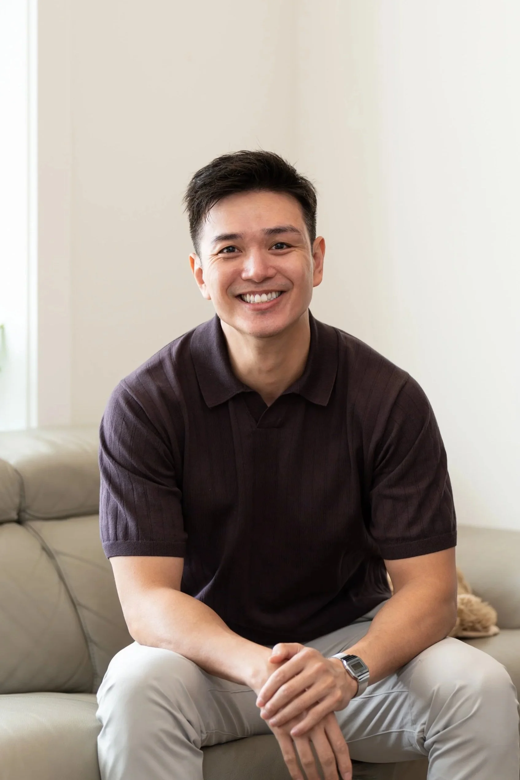 A young man smiling, sitting on a beige sofa in a bright living room, wearing a dark brown polo shirt and light-colored pants, with a large window in the background.