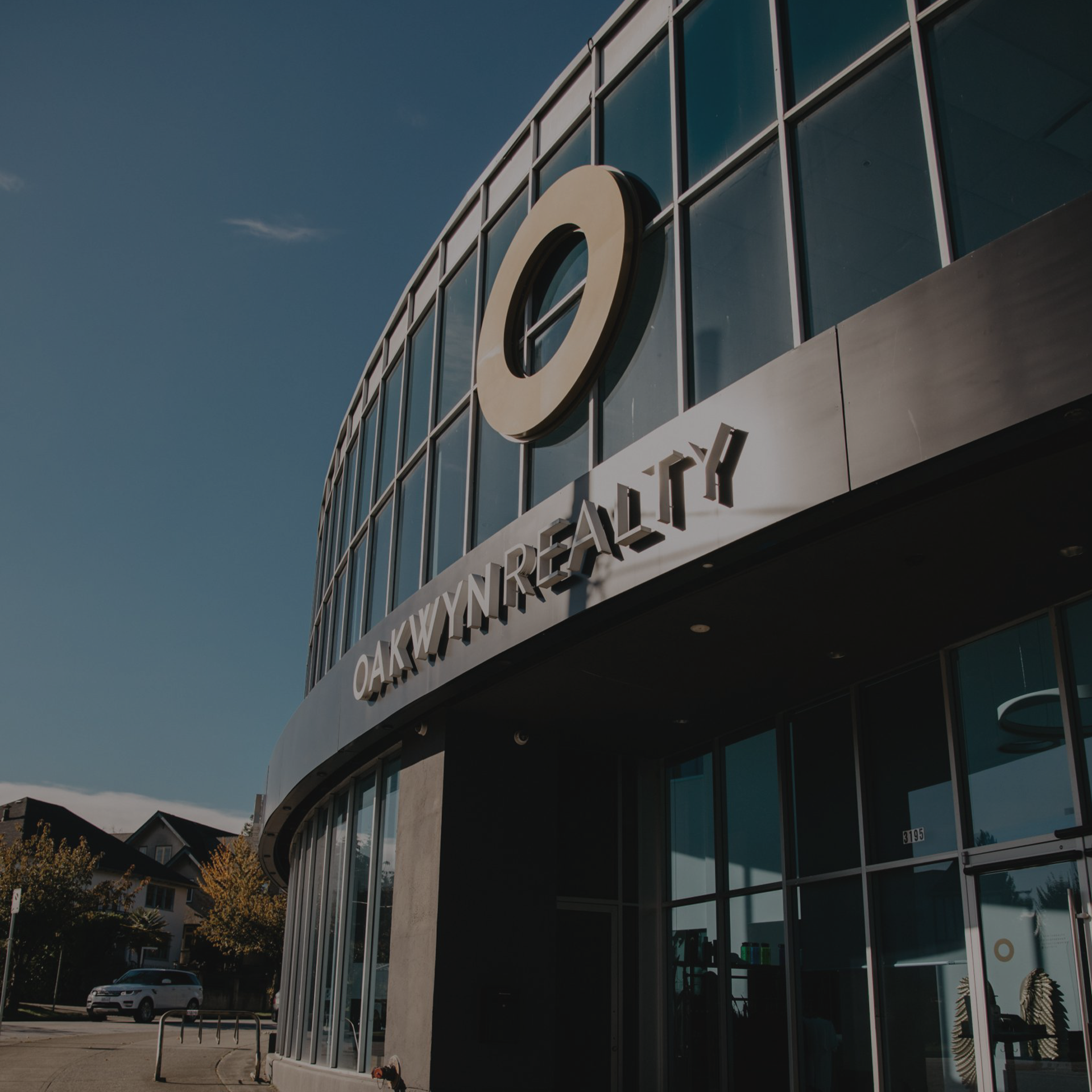 Oakwyn HQ building with large glass windows and a prominent logo on the exterior, with a clear sky in the background and parked cars visible below.