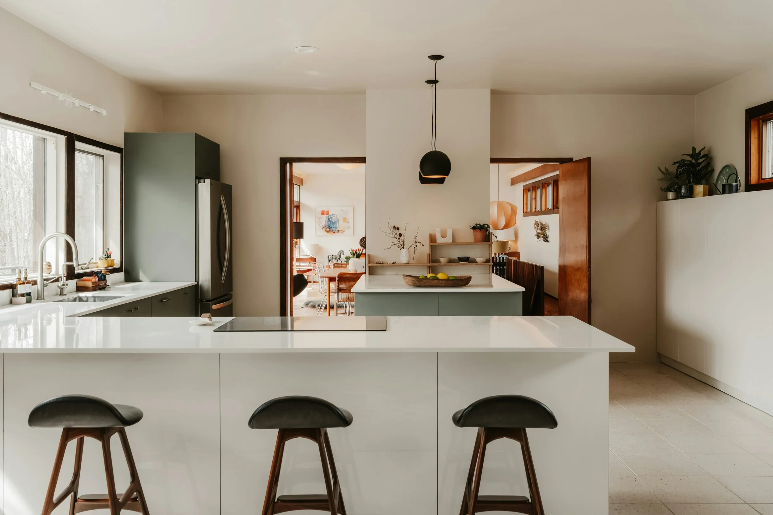 Modern kitchen with white island, black stools, stainless steel refrigerator, and natural light from windows.
