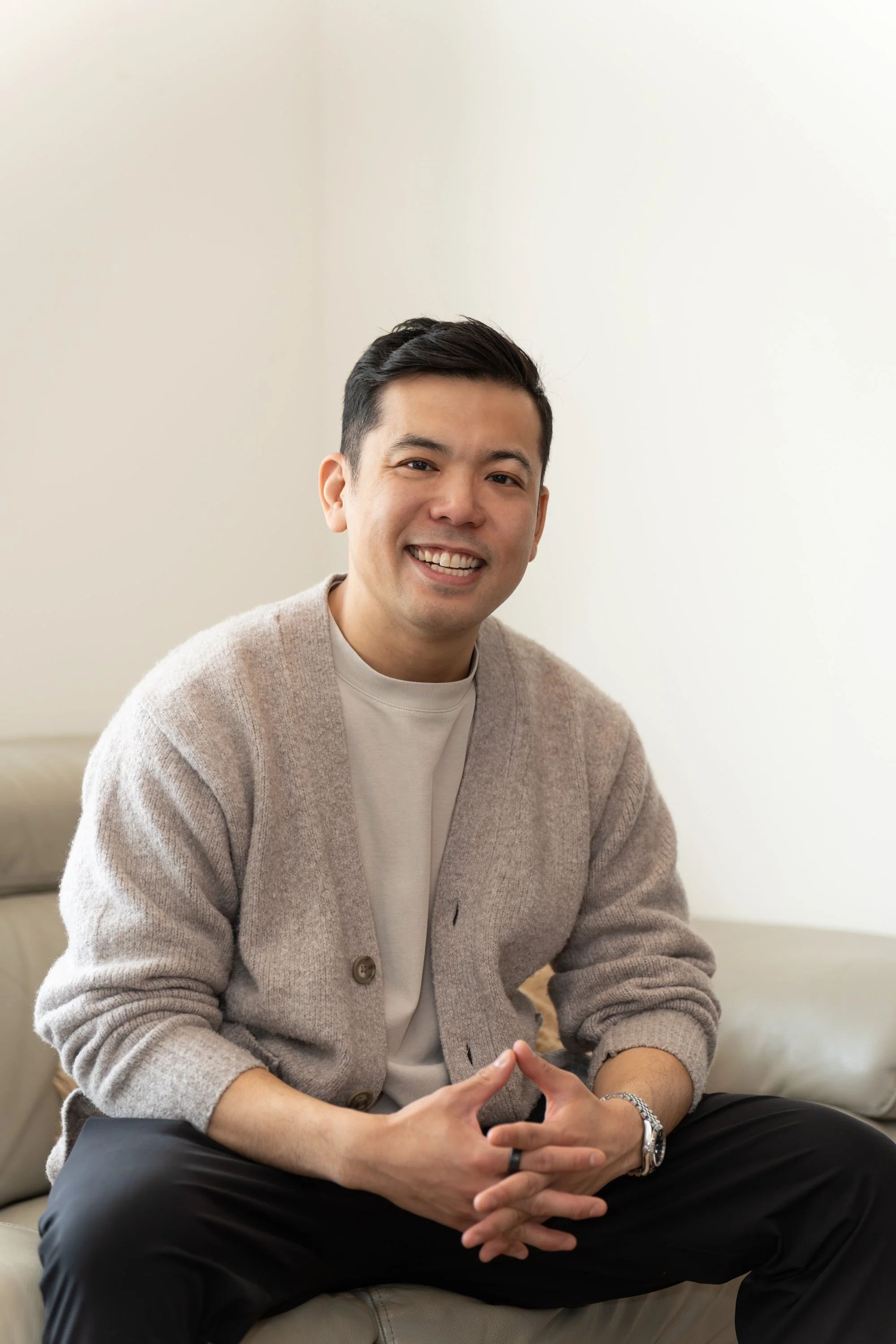 A smiling man with dark hair, wearing a beige cardigan over a white shirt, sitting with hands clasped in front of him against a light-colored wall.
