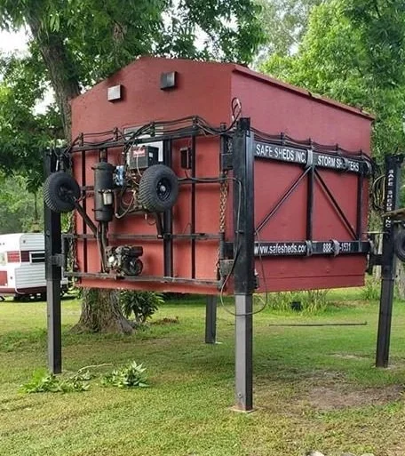 A red shoe storage unit with wheels and straps, elevated on metal supports, with a sign reading 'Safe Shoes Inc.' on the side, located outdoors on a grassy area with trees in the background.