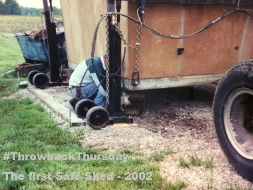 A person working on the underside of a concrete shelter in a rural outdoor setting, with a tractor wheel visible nearby, and a caption reading #ThrowbackThursday The first Safe Shed - 2002.