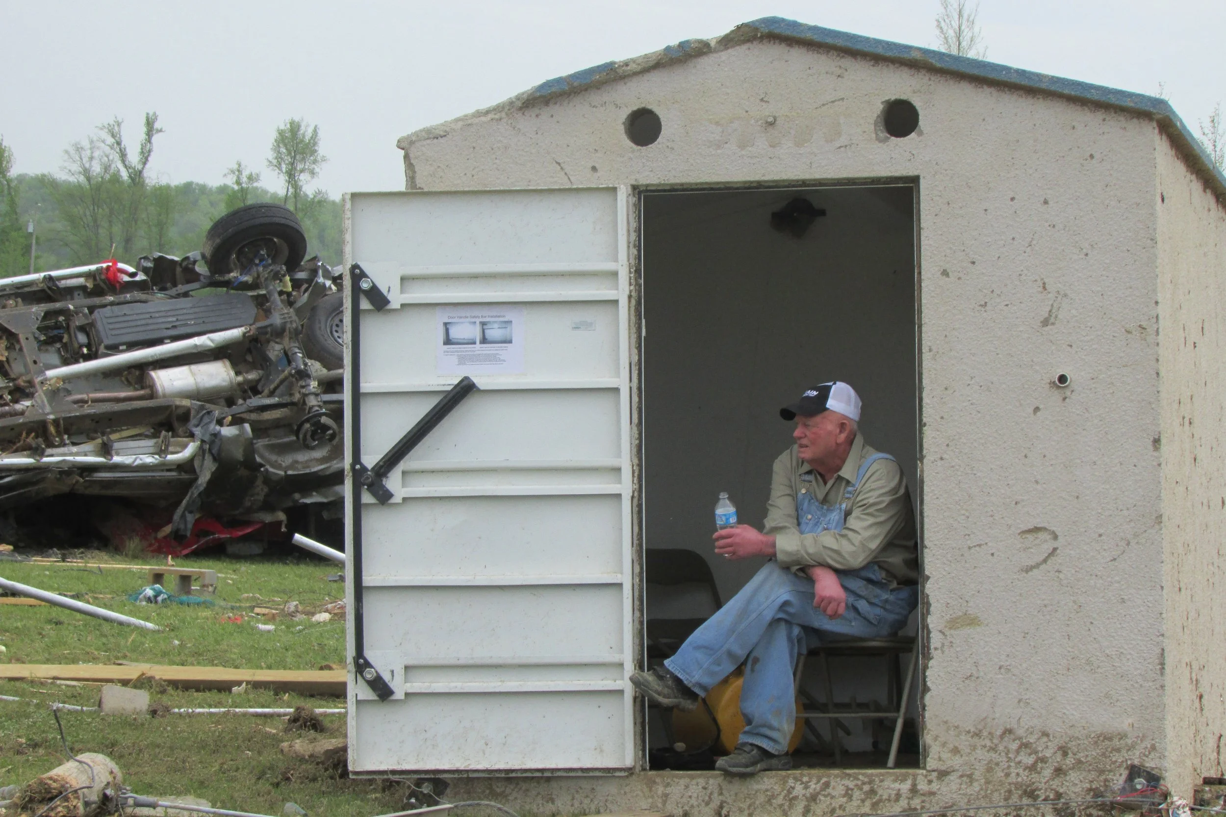 A man sitting outside a small building, holding a water bottle, with a flipped car in the background and debris on the grass.