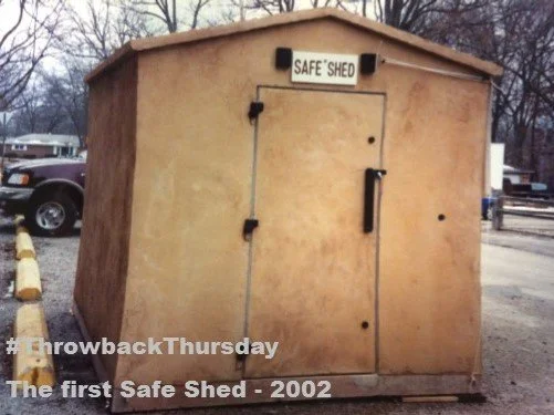 A small, beige concrete shed labeled 'SAFE SHED' with a black handle on the door, located outdoors near parked cars.
