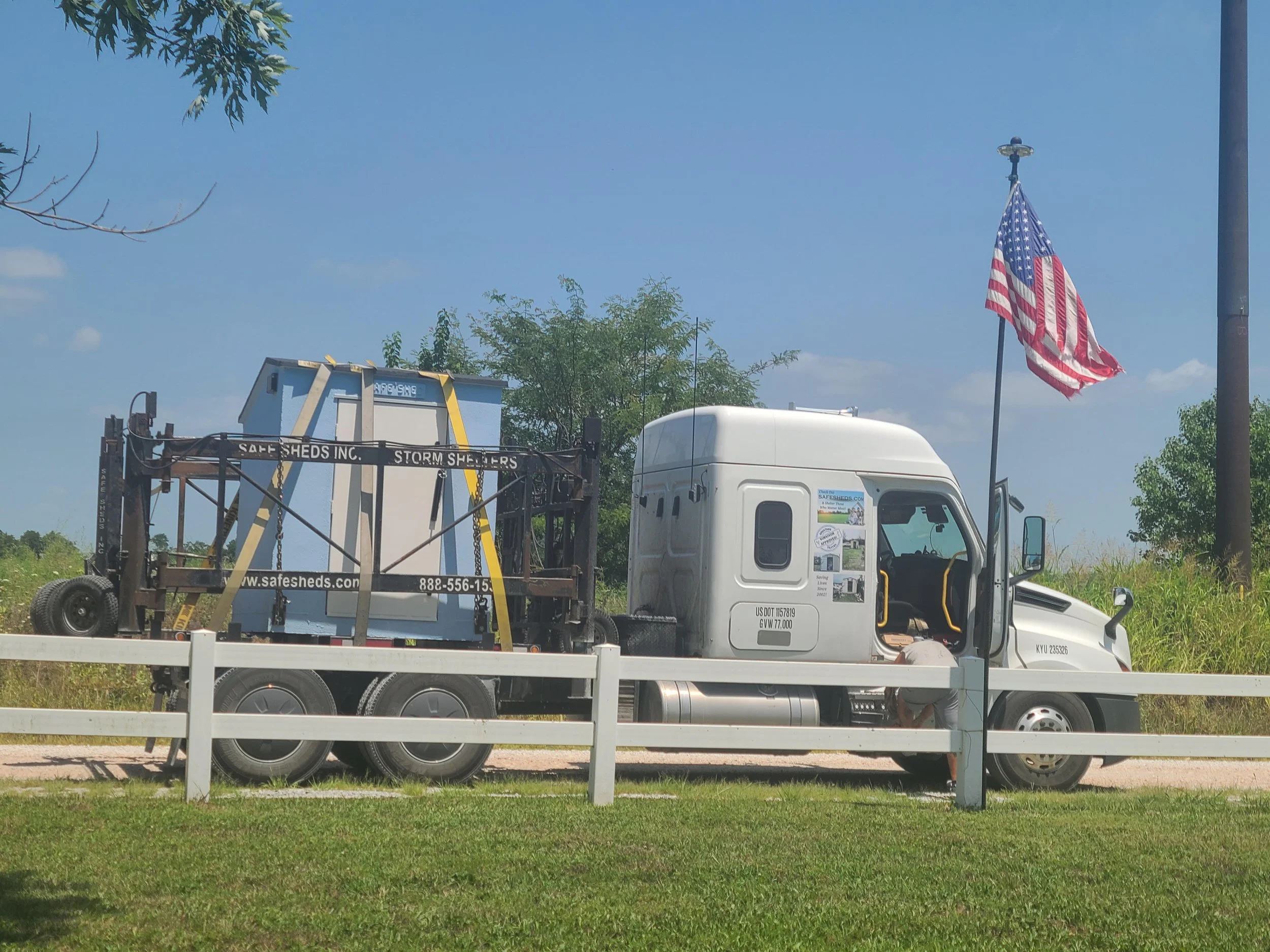 A white truck with a black metal structure on its bed, containing a small, blue, partially enclosed building or unit labeled 'SAFE SHEDS INC.' with a website and phone number, parked near a white fence with a blue sky, trees, and an American flag flying on a pole in the background.