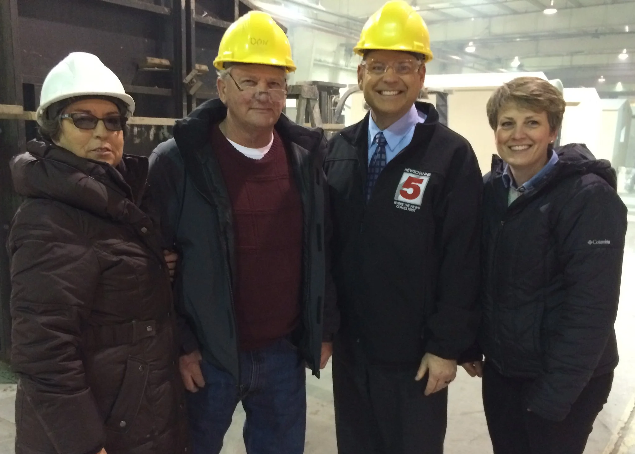 Four people in a factory or industrial setting, wearing safety helmets and jackets. Two men are wearing yellow helmets, and one woman is wearing a white helmet. They are smiling and standing close together.