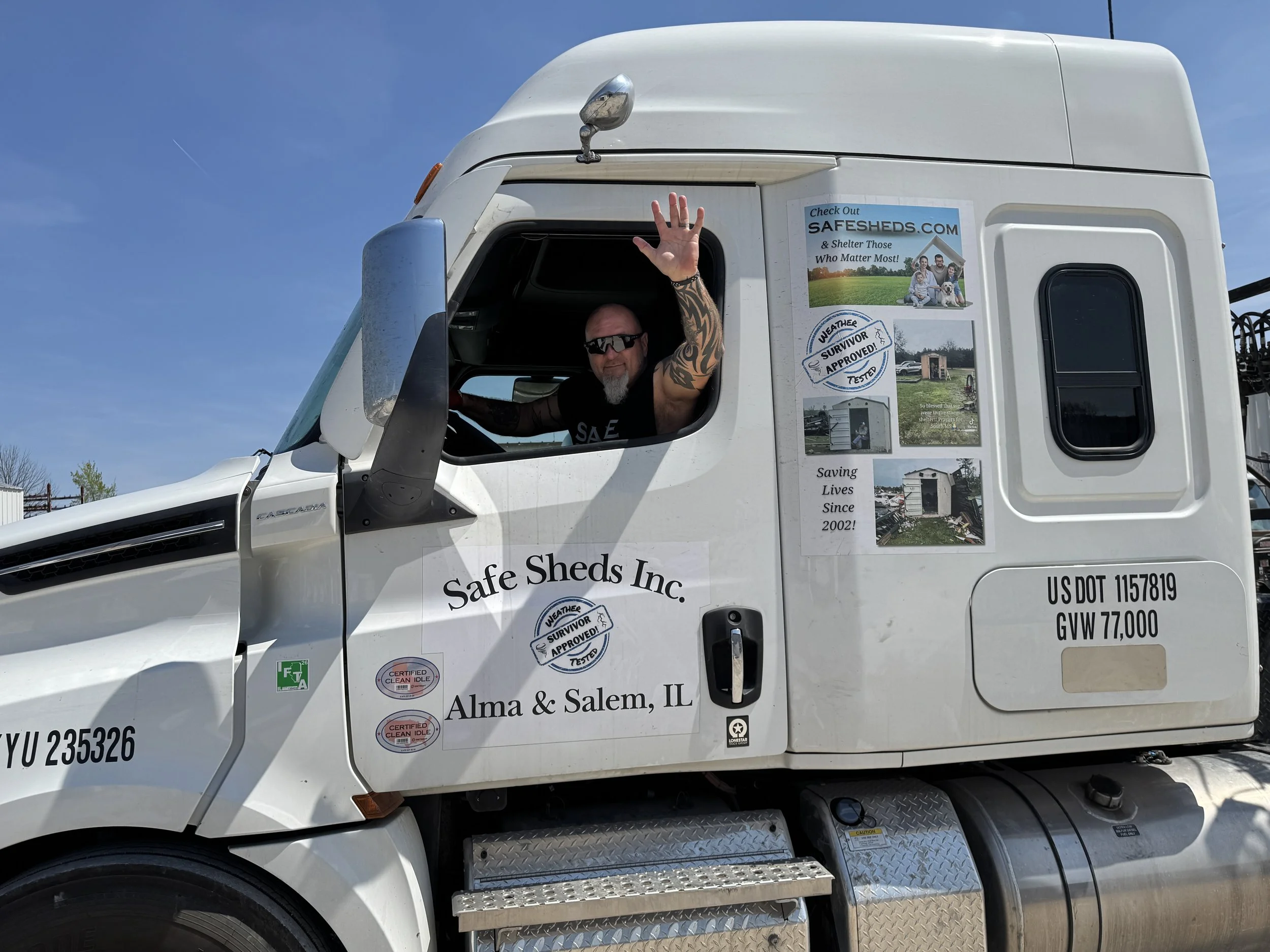 A white semi-truck with promotional stickers on the side for Safe Sheds Inc., a shed business in Alma and Salem, Illinois. A man with tattoos and sunglasses is waving from the open window of the truck's cab, with a clear blue sky in the background.