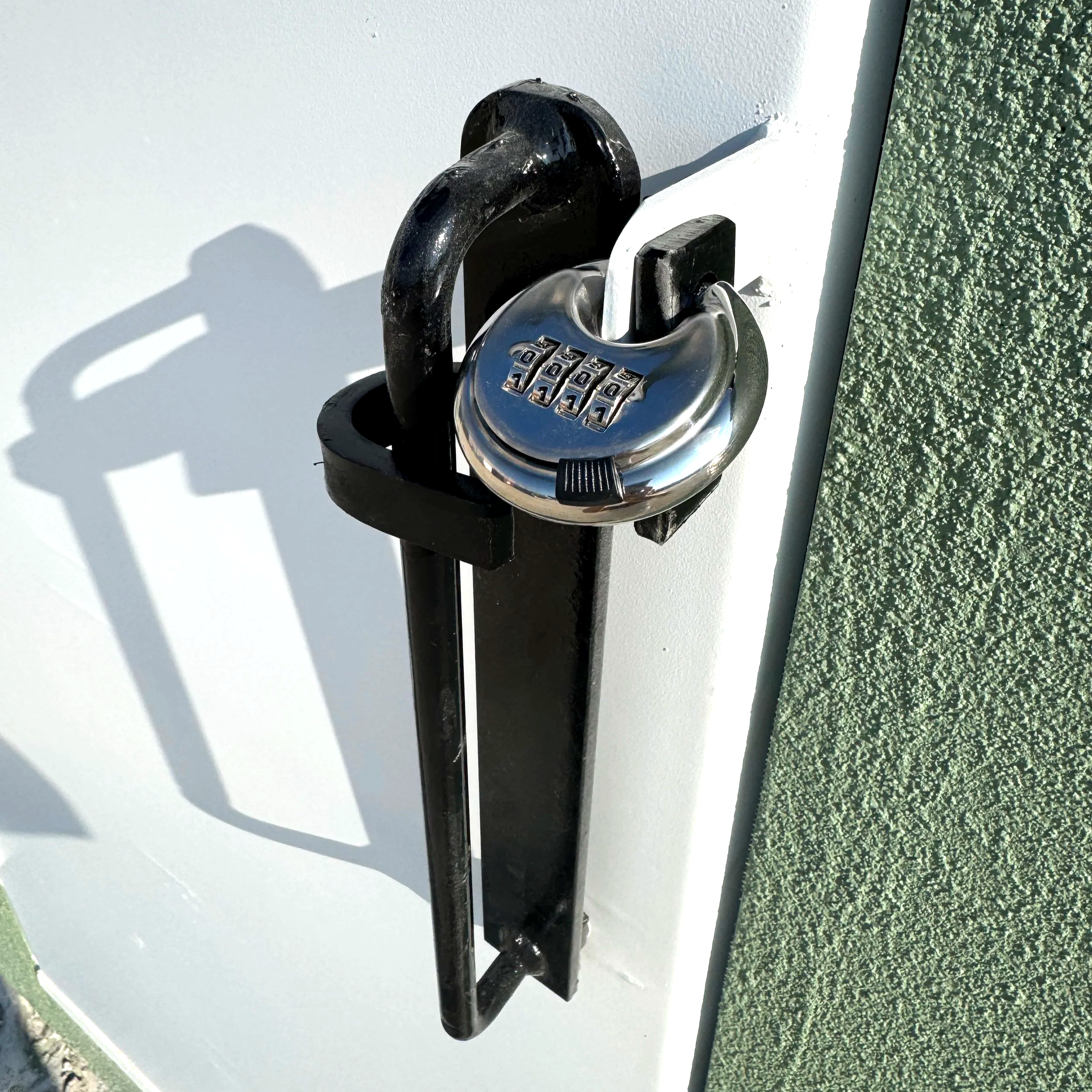 Close-up of a black combination padlock attached to a white surface with textured green wall on the right side, and shadows cast by the lock and shackle.