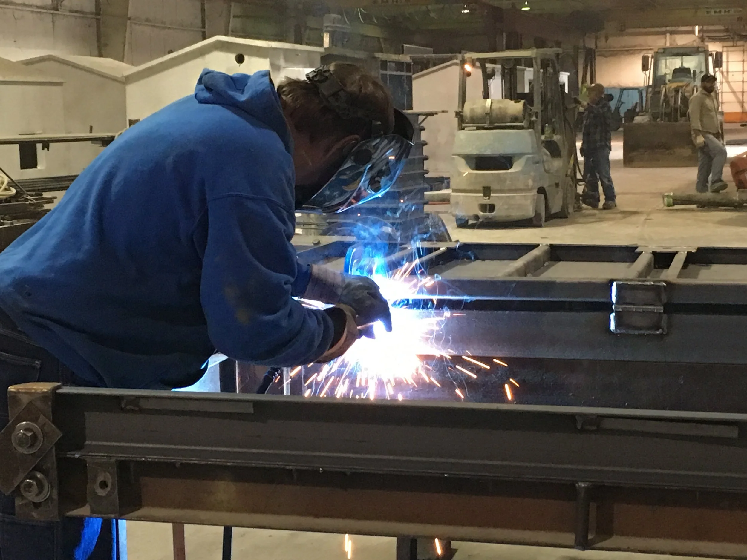 Worker welding metal parts in an industrial workshop with several machinery and workers in the background.