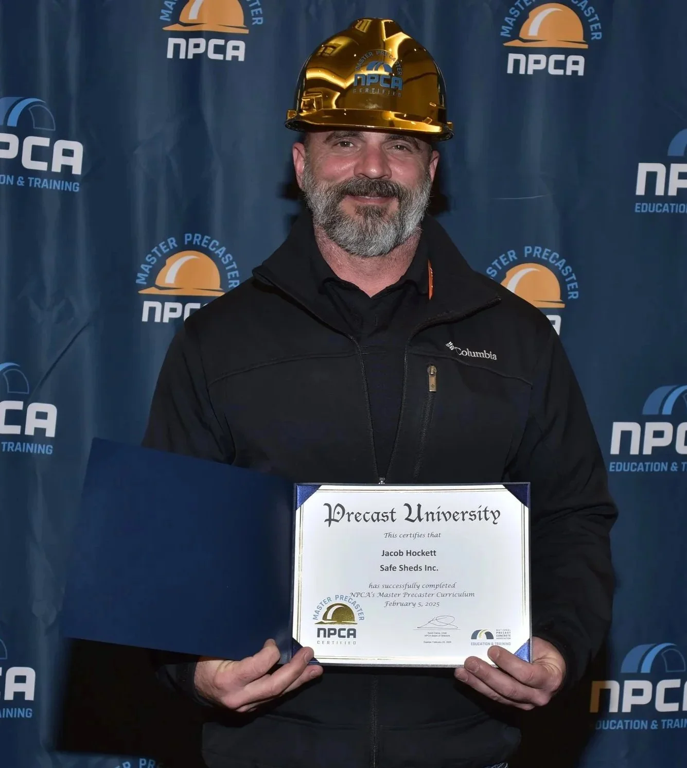 A man with a gray beard wearing a yellow hard hat and black Columbia jacket, smiling, holding a framed certificate that reads 'Precast University' awarded to Jacob Hockett, in front of a blue backdrop with logos of NPCA.