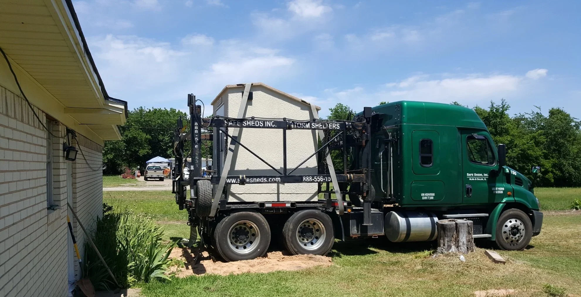 A green truck with a lift platform is parked outside a building, lifting a small white shed towards the roof.
