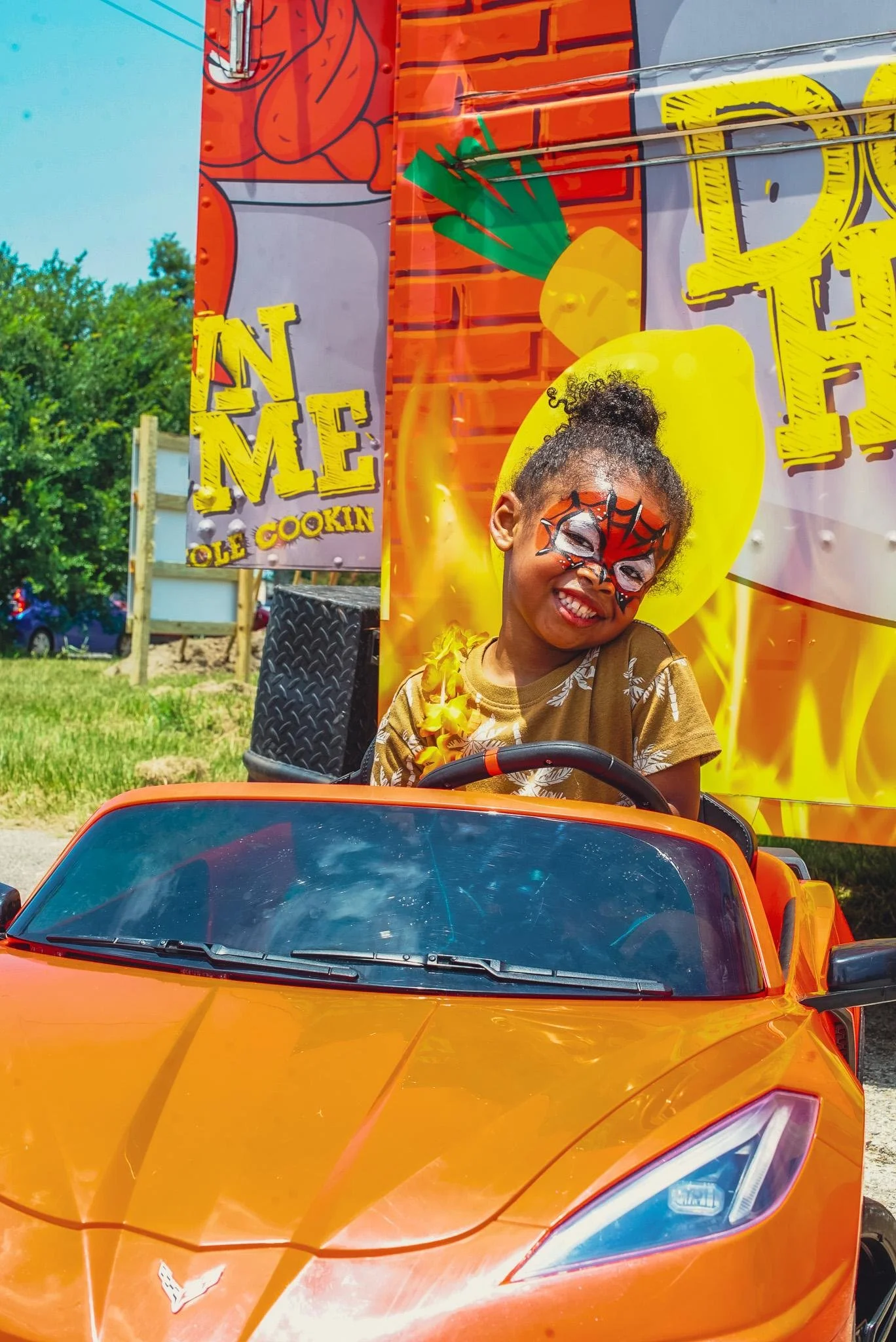 Young girl with face paint sits in a motorized toy car and smiles for the camera at Toddler’s University.