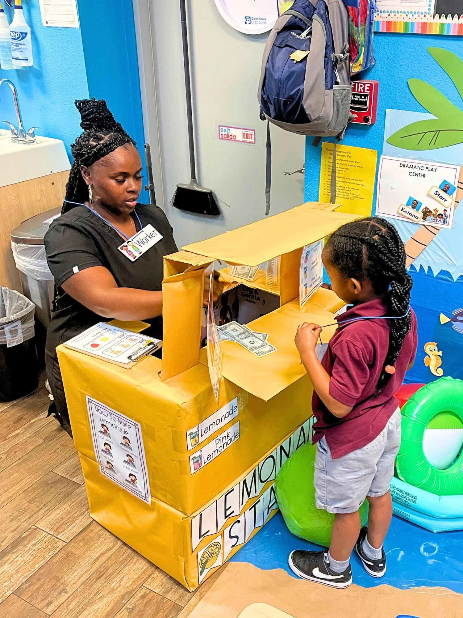 Young girl and teacher participate in a lemonade stand activity, with the student practicing spending money while the teacher manages the stand.