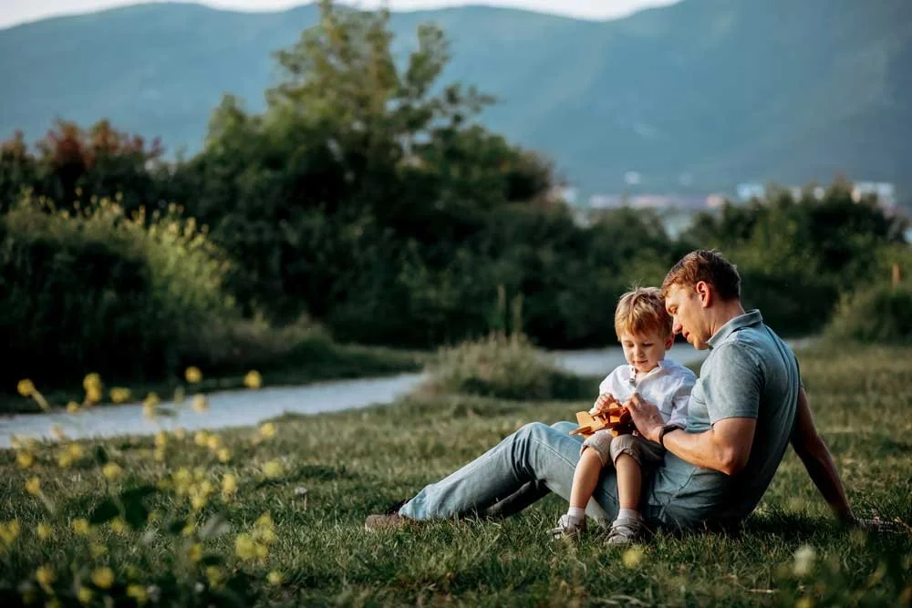A father and young son sitting on grass near a river, looking at a book together, in a lush, scenic outdoor setting.