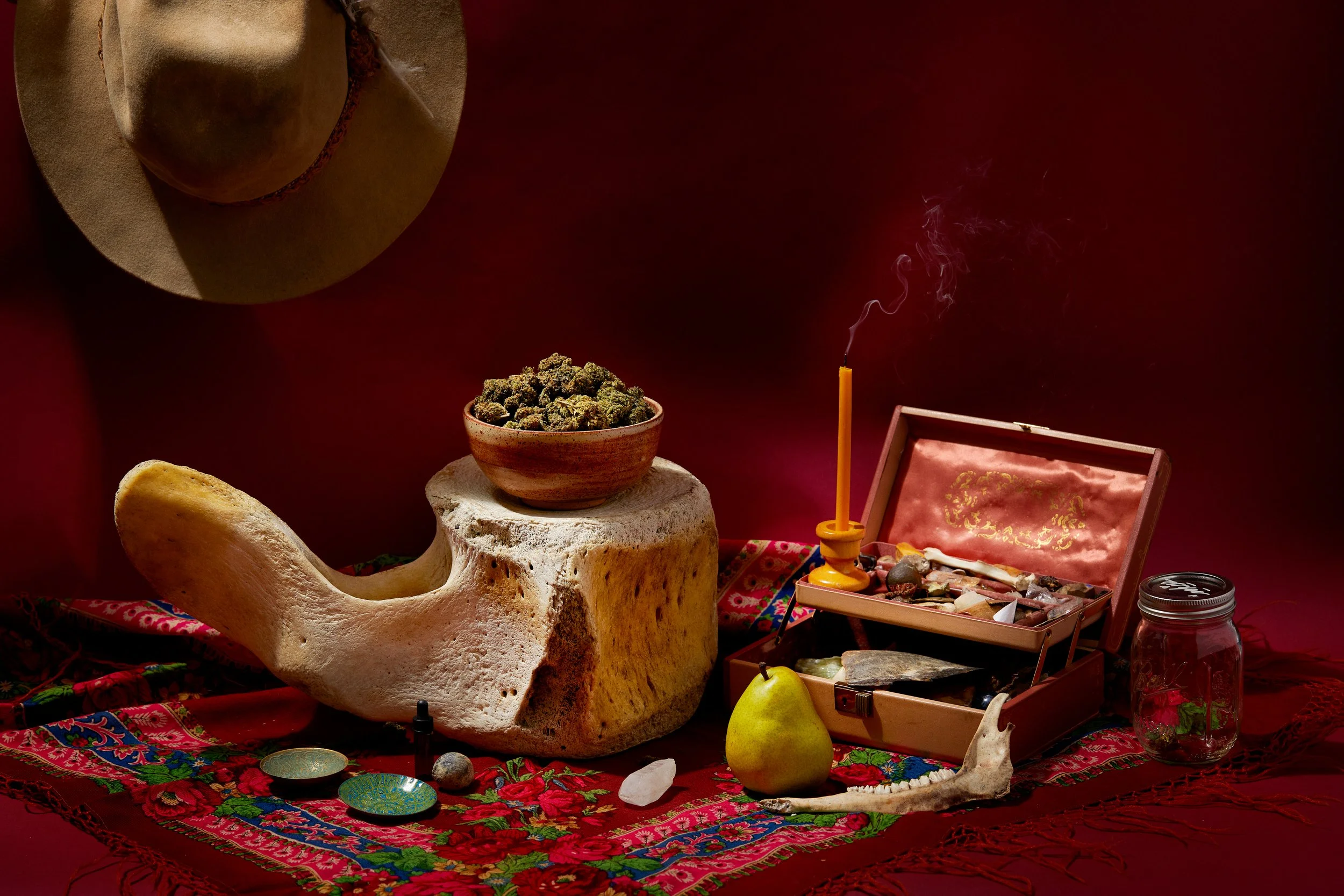 A collection of items on a colorful patterned cloth, including a hat, a bowl of cannabis buds, a lit incense stick, a jewelry box with stones, a pear, a white animal skull, and a jar of small stones, with a dark red background.