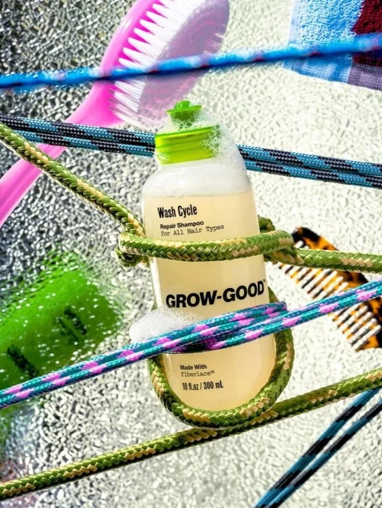 A bottle of Grow-Good Wash Cycle Repair Shampoo hanging on a colorful clothesline with a shower brush, a green sponge, and a butterfly in a bright outdoor setting.