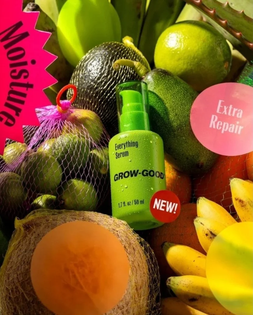 A collection of fresh fruits including oranges, bananas, limes, and an avocado, with a green skincare serum called GROW-GOOD Everything Serum in the center. Labels highlight features like 'Moisturize' and 'Extra Repair.'