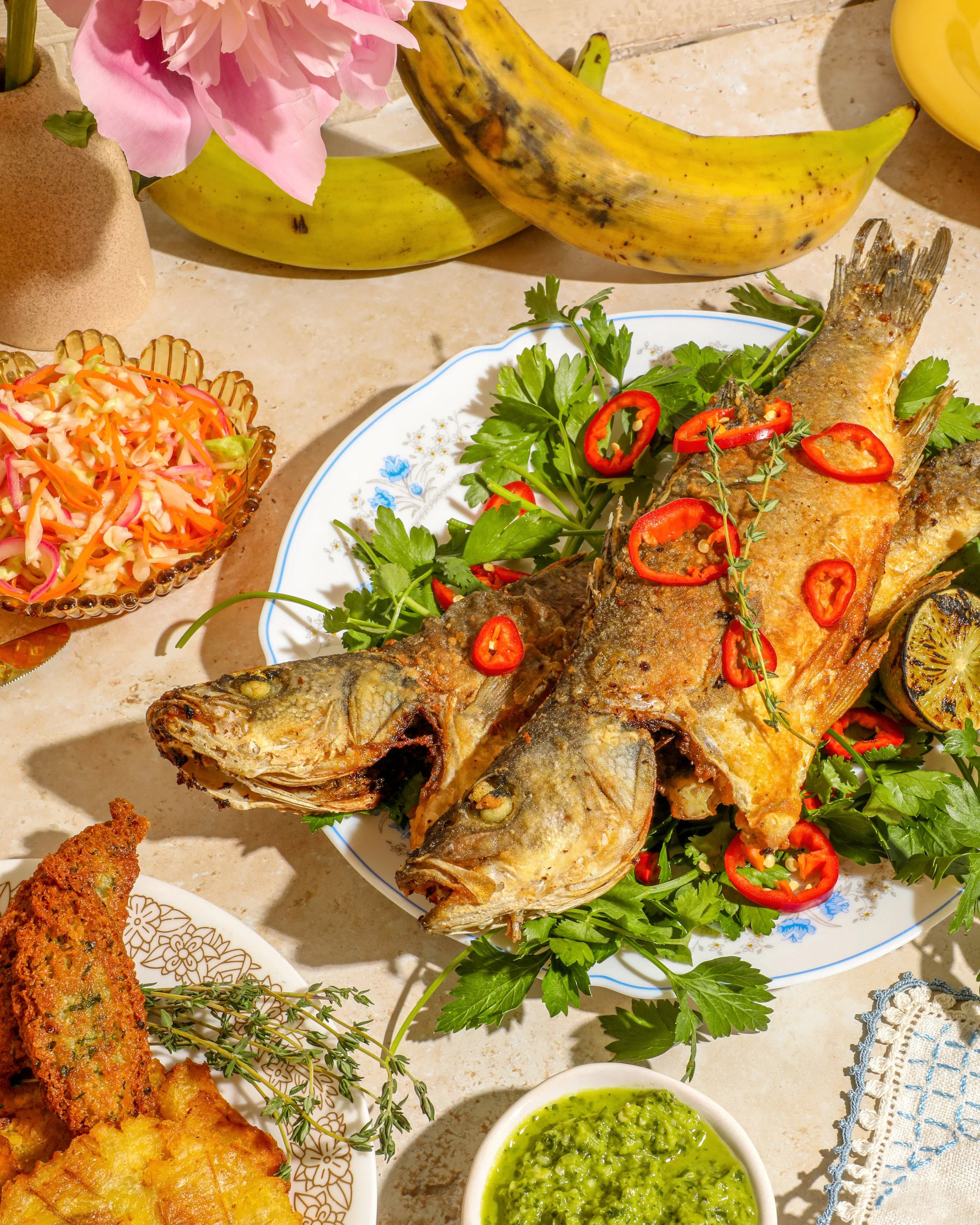 Fried fish garnished with red chili slices and fresh herbs, served on a white plate with parsley and a lemon wedge, surrounded by various dishes including coleslaw, green dipping sauce, and bananas, on a kitchen countertop.