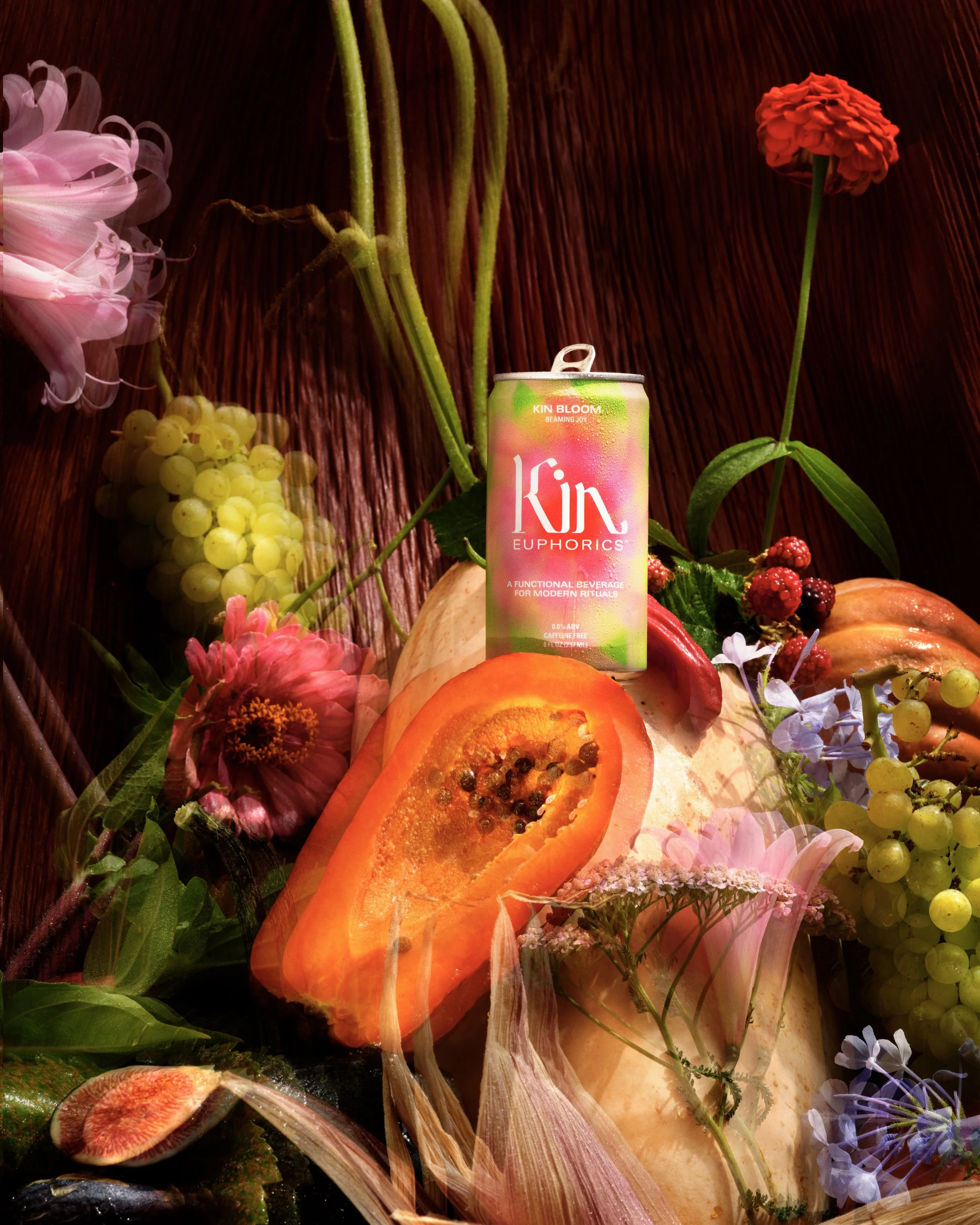 A can of Kin Euphorics drink surrounded by various flowers and fruits, including a papaya, red berries, white grapes, and pink blossoms, arranged on a wooden surface.