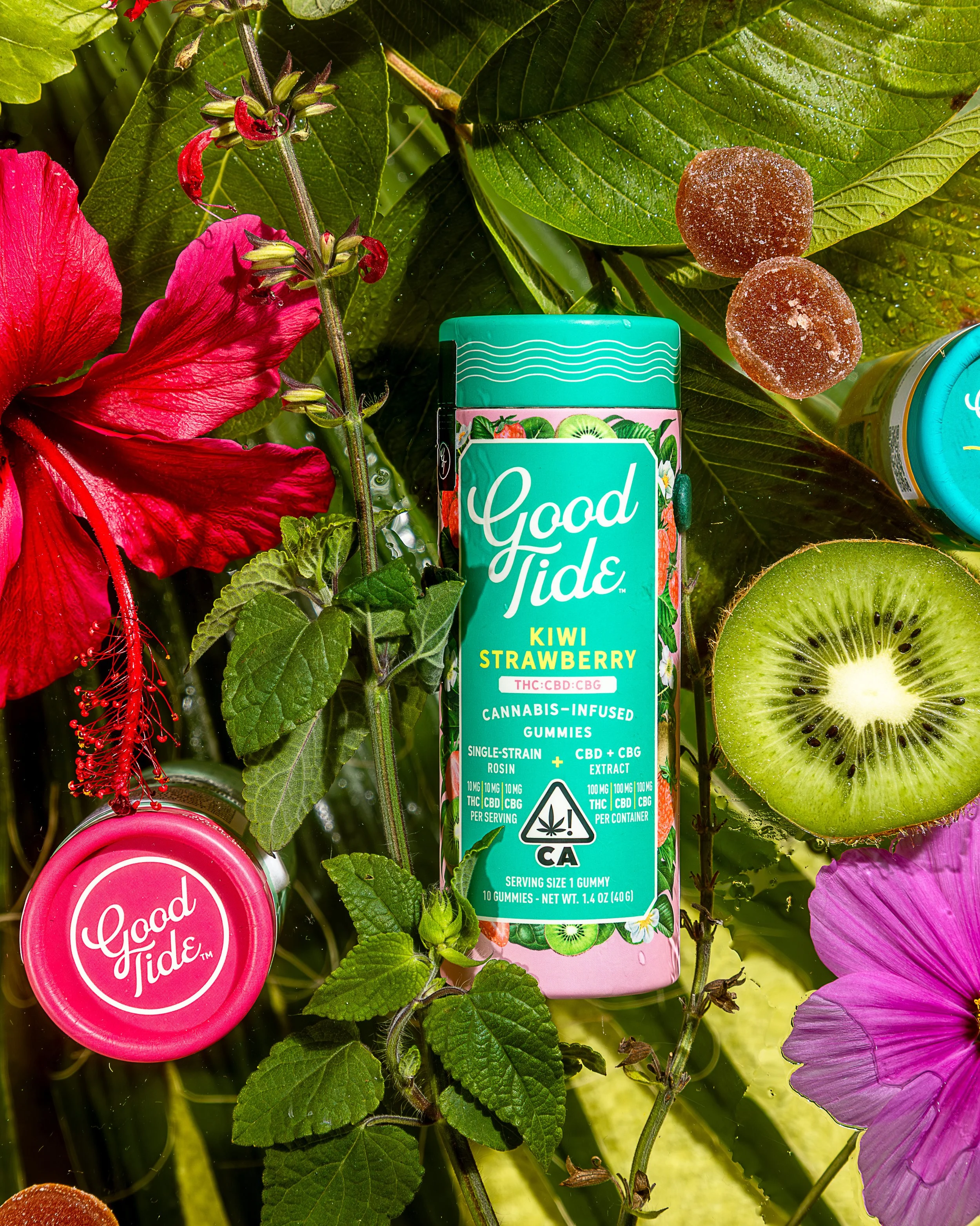 A cannabis-infused gummy container labeled 'Good Tides Kiwi Strawberry' surrounded by pink hibiscus flowers, kiwi, lemon, and green leaves.