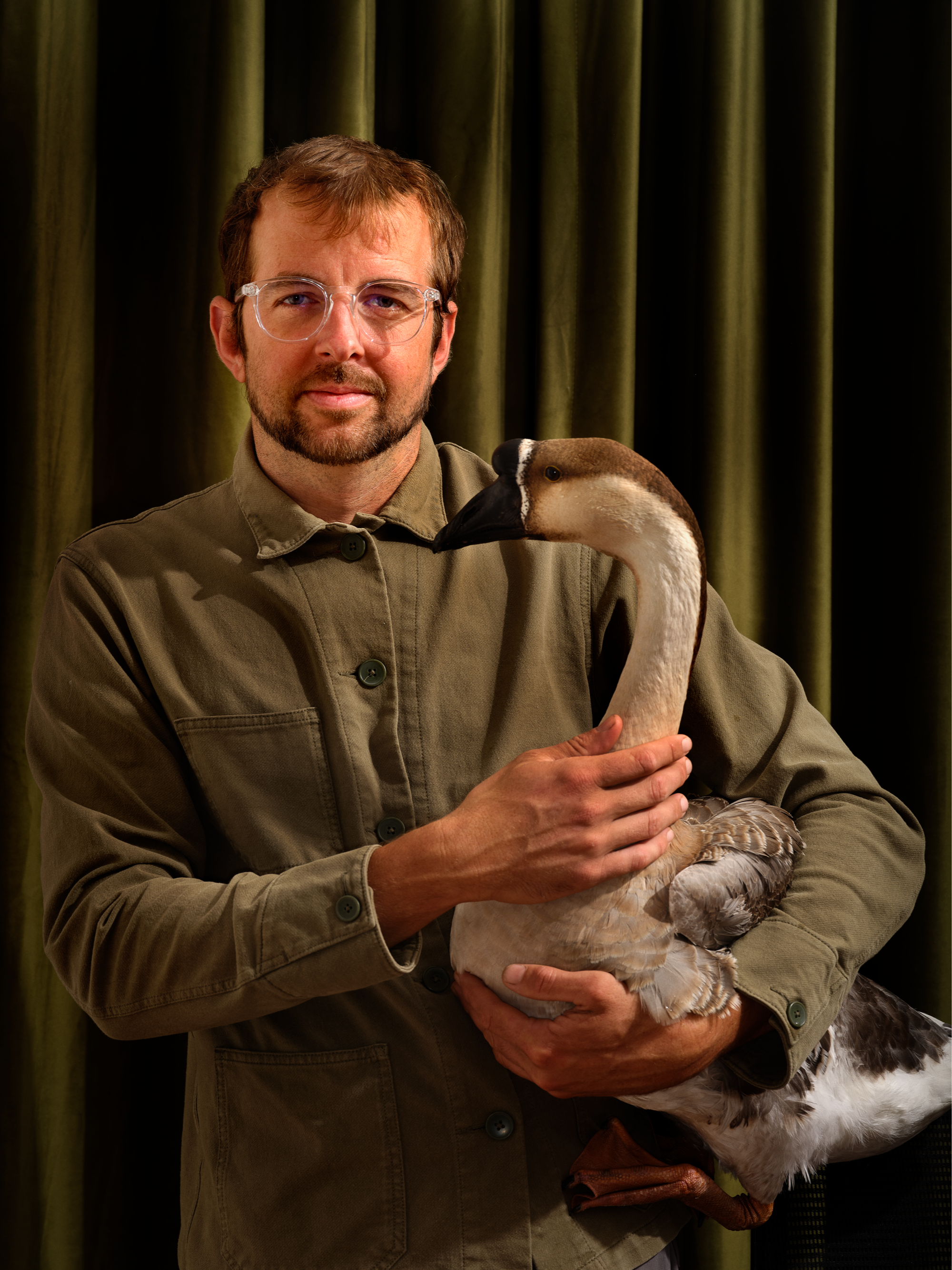 Man with glasses holding a goose in front of green curtains.