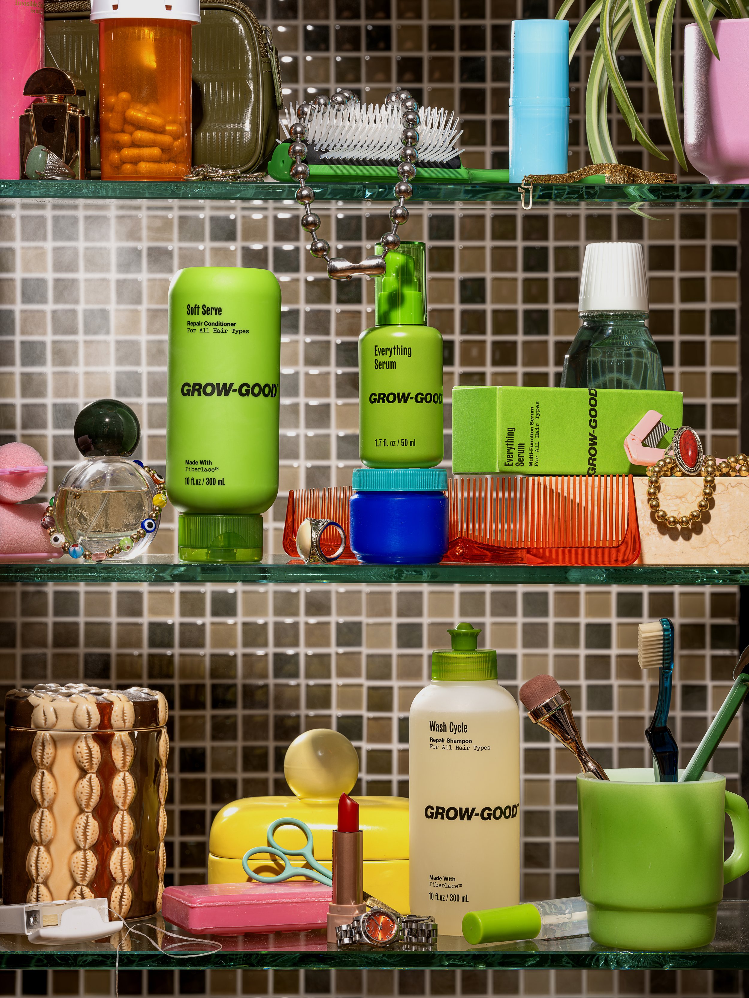 Organized bathroom shelf with hair care products, jewelry, and toiletries. Green bottle labeled 'Grow-Good Everything Serum' and other colorful containers. Jewelry including a beaded necklace and rings, hairbrush, and bathroom accessories are also vi