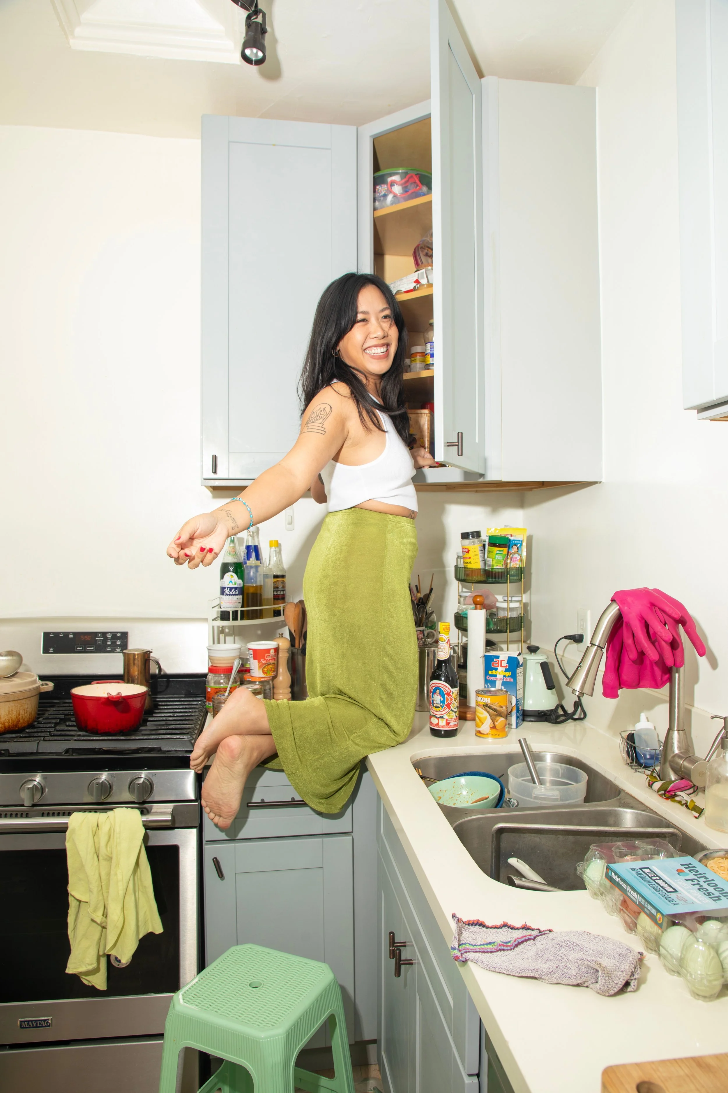 A woman with long dark hair kneeling on a kitchen counter, smiling and pointing at the camera. The kitchen has light gray cabinets, a stove, and various kitchen items on the countertop, including bottles, bowls, and containers.