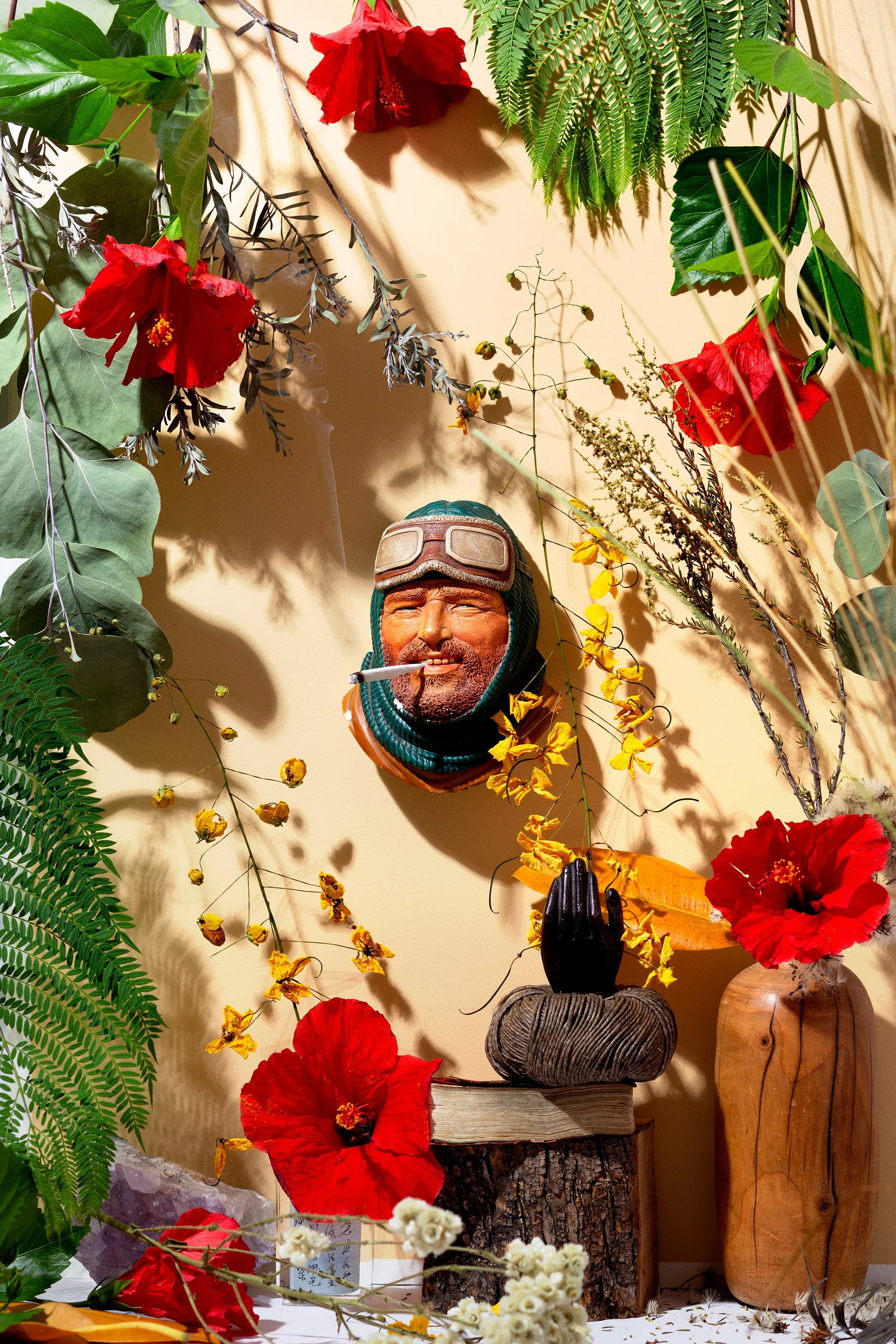 Decorative display featuring a wooden career, lush green leaves, red hibiscus flowers, yellow flowers, a ceramic head with goggles, a pile of wood, and ornamental crystals.