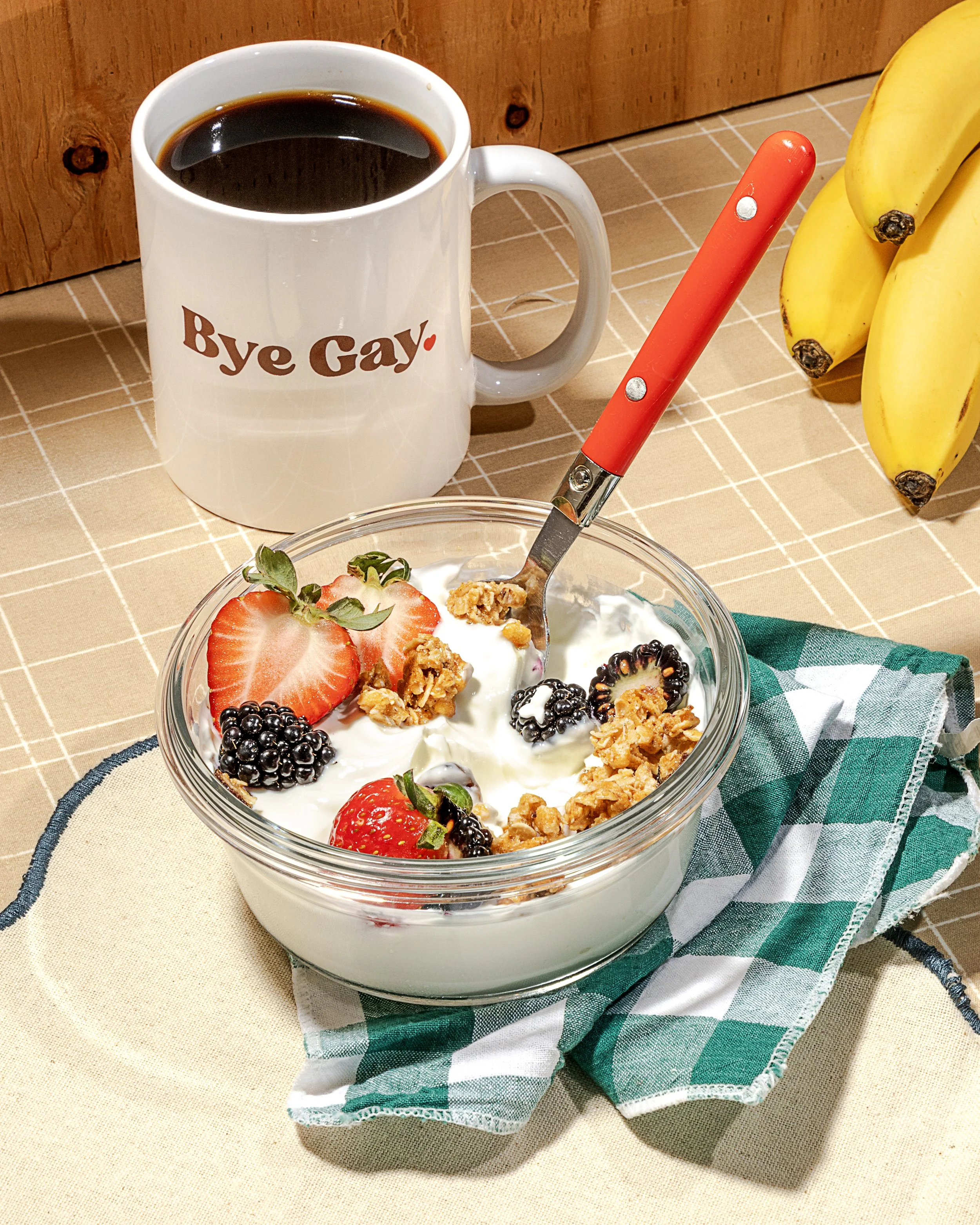 A white mug with the words "Bye Gay" printed on it, filled with black coffee, sitting on a beige checkered tablecloth. To the right of the mug, there are bananas, and in the foreground, a bowl of yogurt topped with strawberries, blackberries, granola