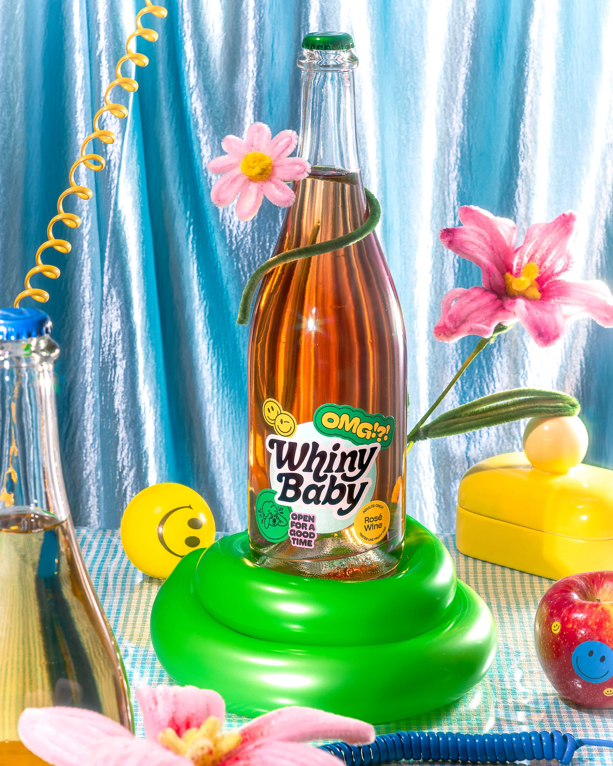 A bottle of rosé wine with colorful stickers and the label 'Whiny Baby', decorated with pink flowers and green vines. Surrounding objects include a smiley face ball, a yellow container, balloons, and a shiny metallic object, all set against a blue cu