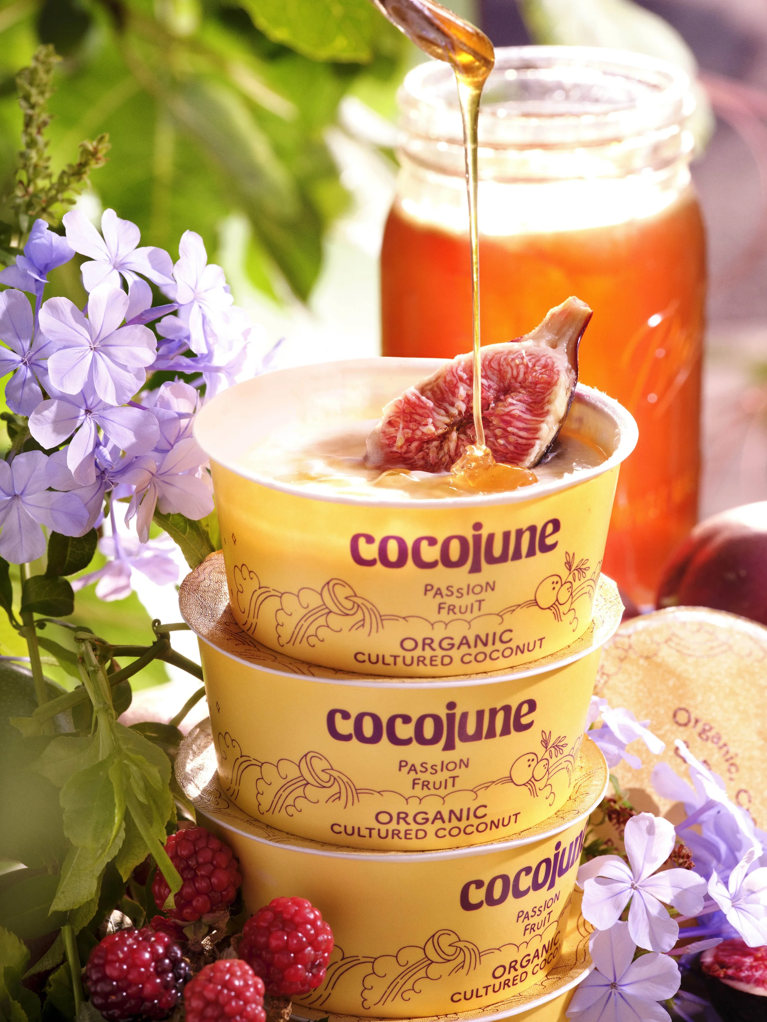 Multiple yellow cups labeled 'CocoJune Passion Fruit Organic Cultured Coconut' with a scoop of pinkish passion fruit pudding and a fruit topping, surrounded by purple flowers and blackberries under natural light.