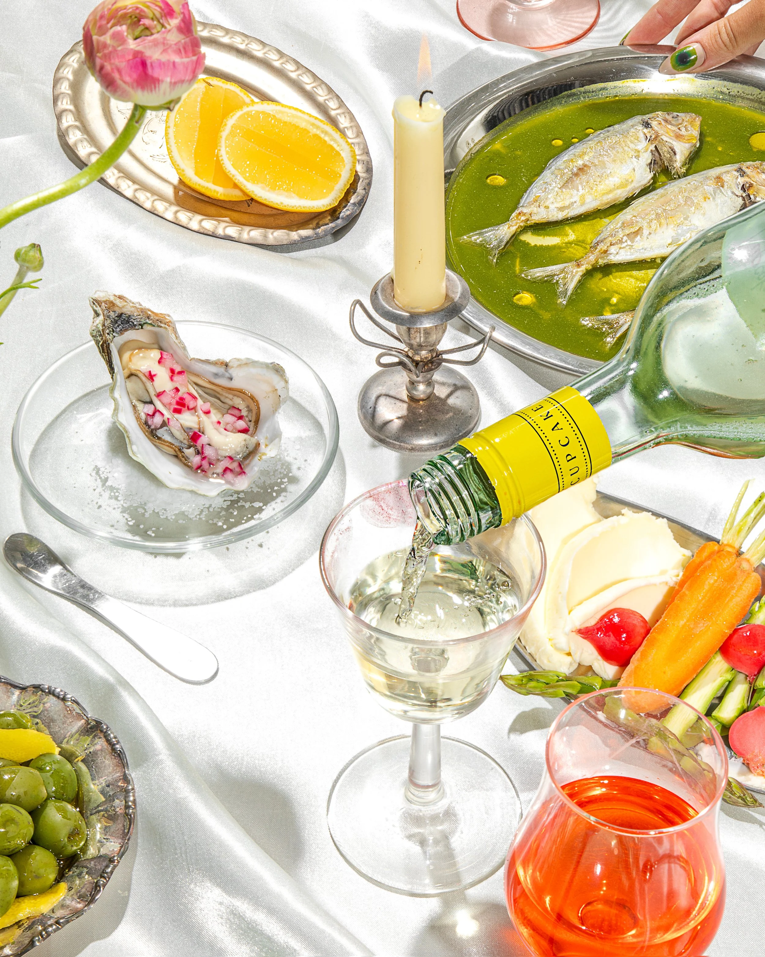 Table setting with seafood, fruit, and beverages, including oysters on a plate, lemon slices, a green fish in oil, green grapes, a pink candle, a glass of champagne, and a bottle of wine, all on a white tablecloth. Drink Styling.