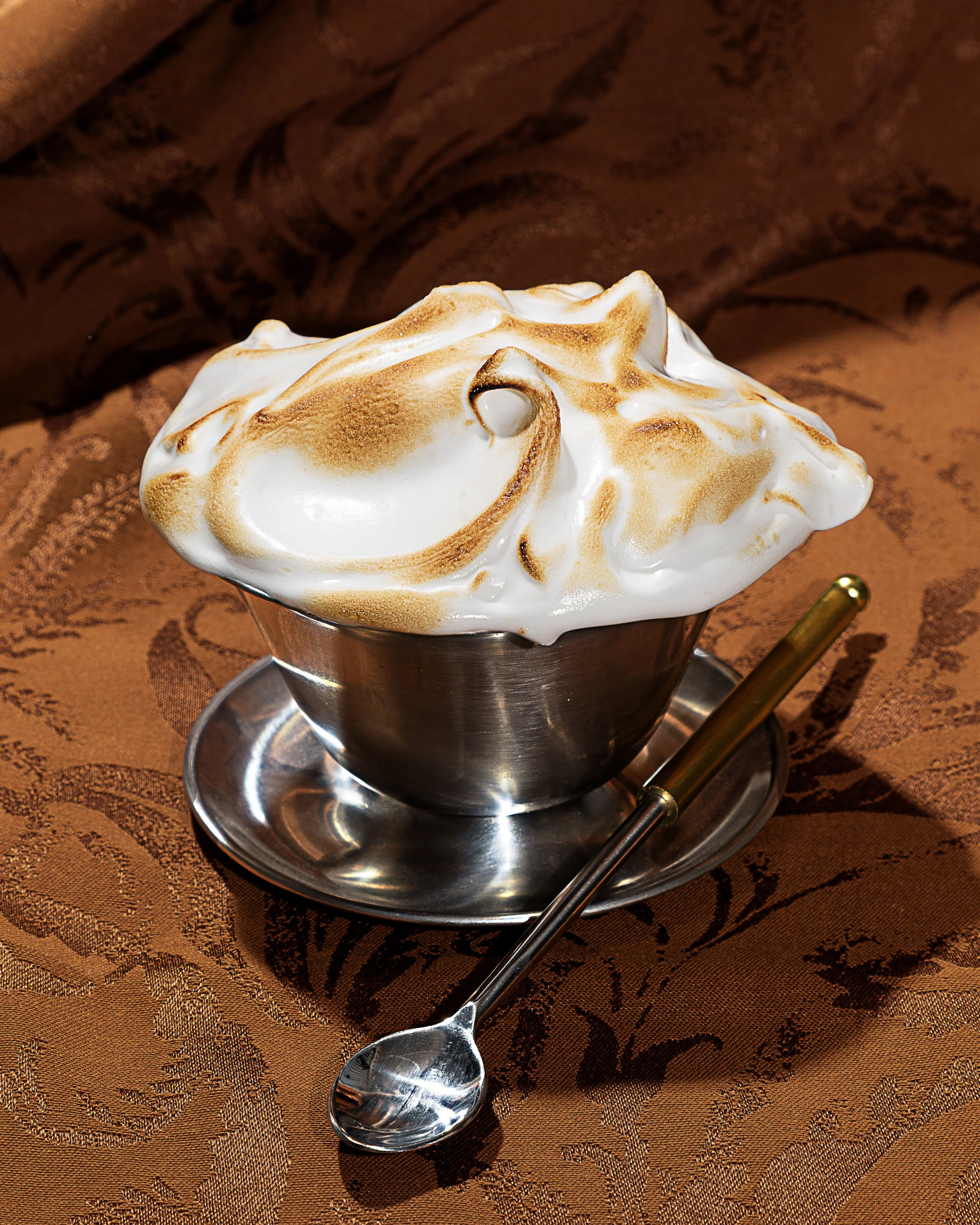 A metal mug filled with a toasted meringue topped with peaks of golden-brown toasted meringue, placed on a textured bronze-colored tablecloth with a pattern, accompanied by a long-handled spoon.