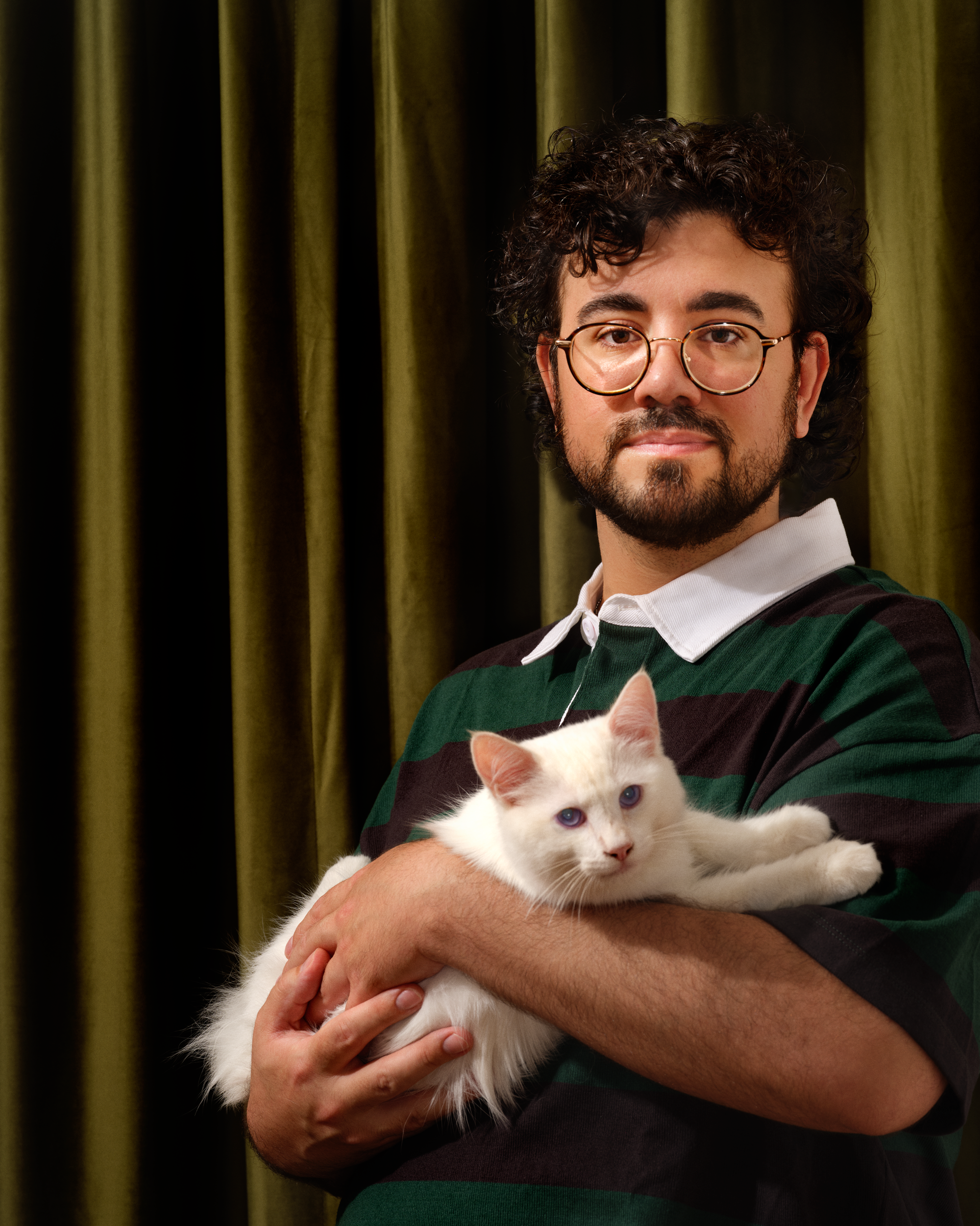 A young man with curly dark hair, glasses, and a beard holding a white cat with blue eyes against a background of green curtains.