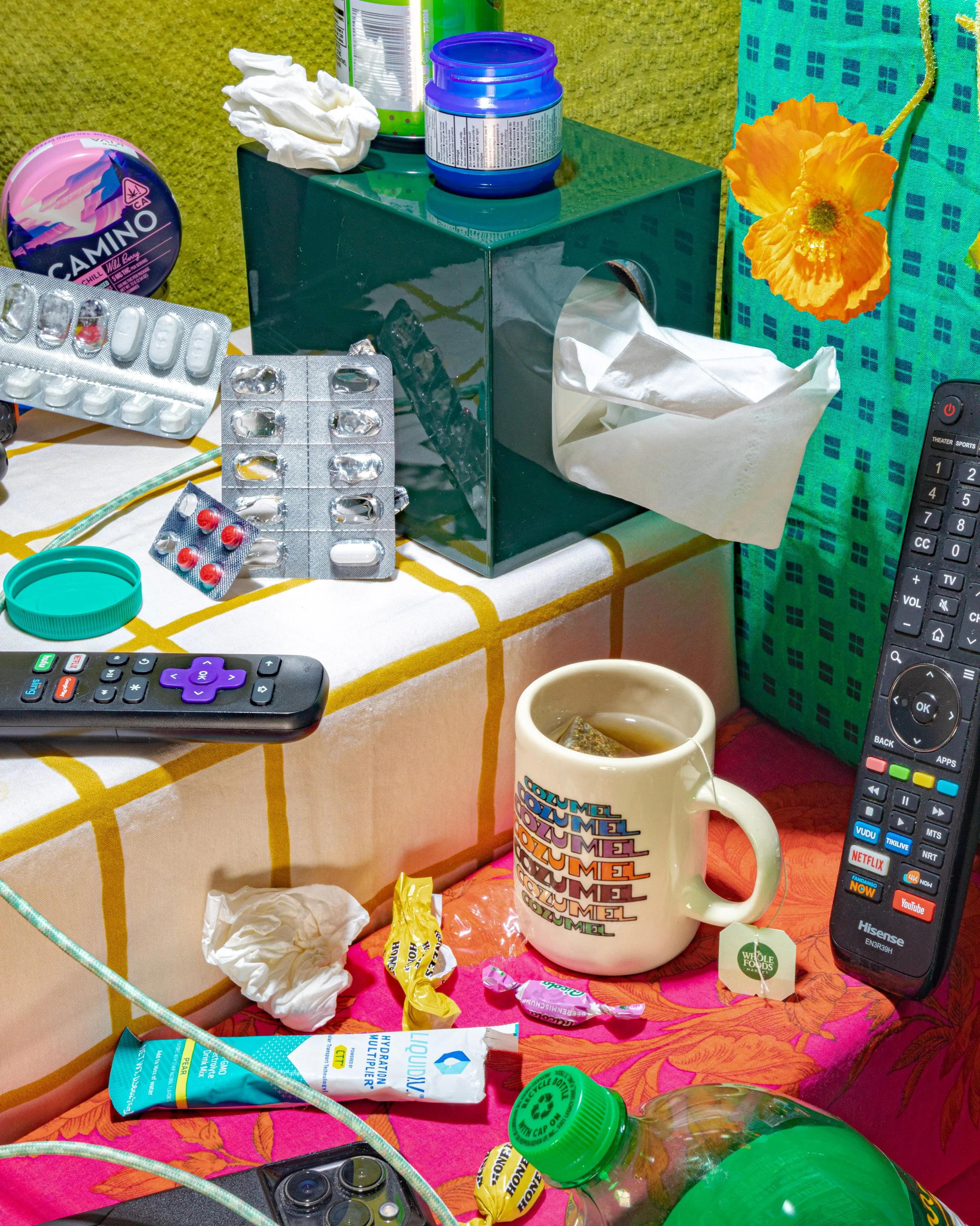 A cluttered table with various items including a tissue box, pill bottles and blister packs, a mug with colorful text, a remote control, a water bottle, a packet of hydration powder, and discarded tissues, set against a green and patterned background