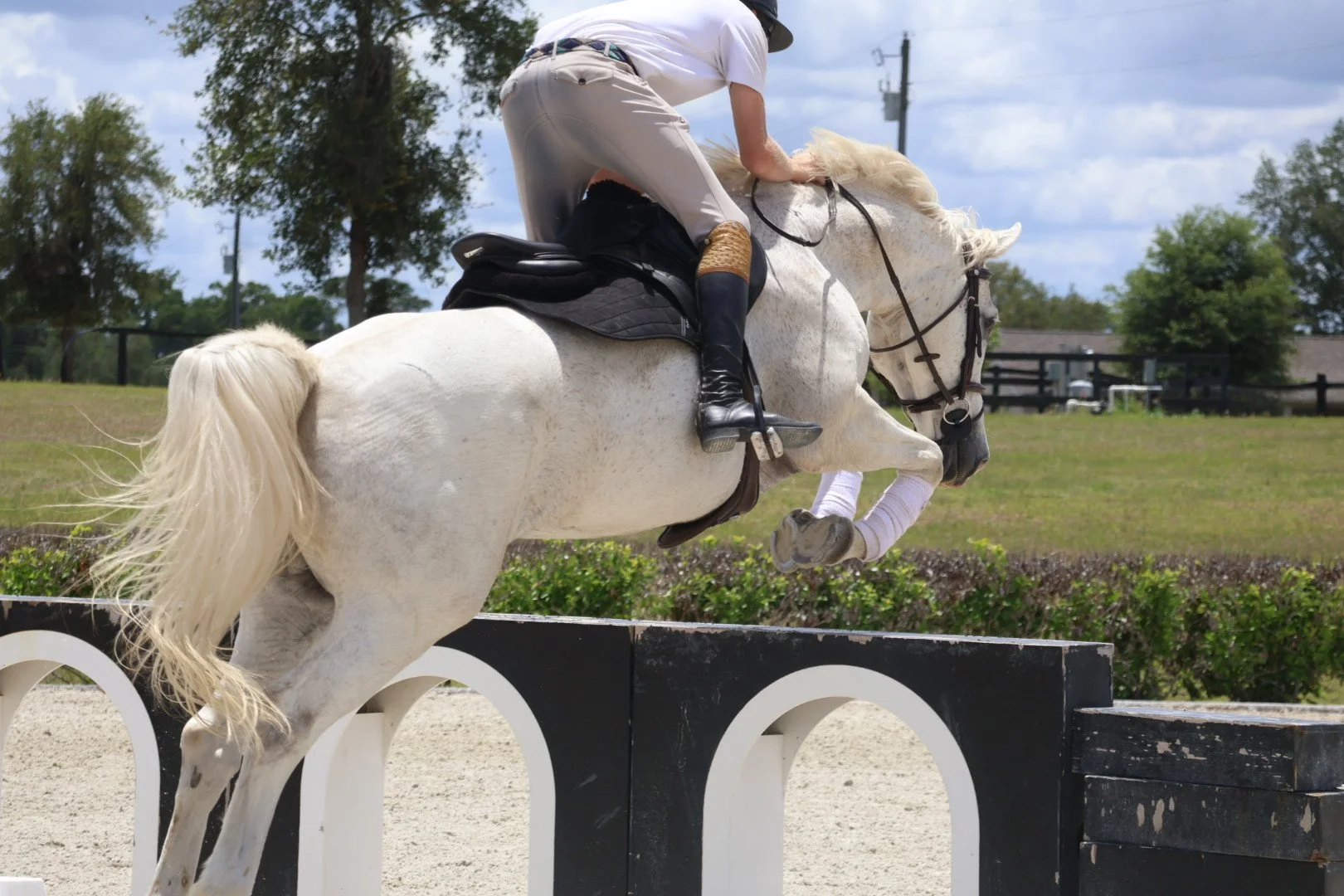 A rider on a white horse jumping over an obstacle on a sandy equestrian arena outdoors during daytime.
