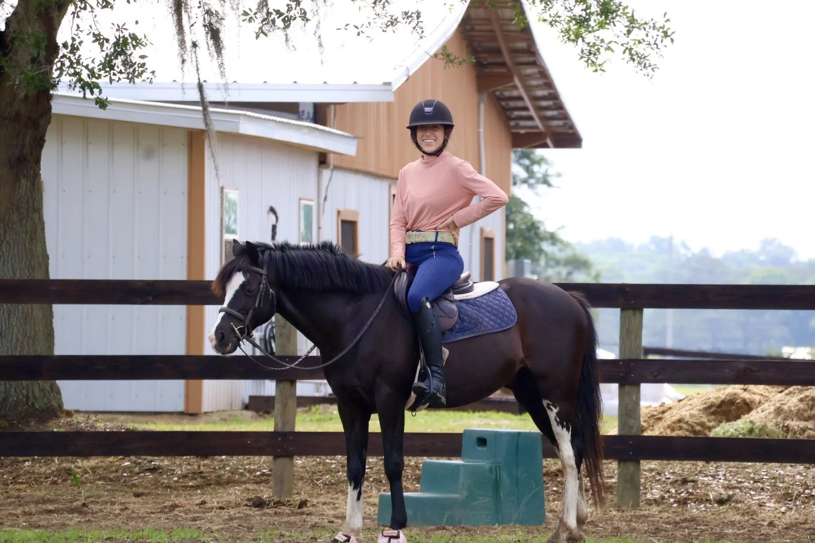 A woman riding a black and white horse in a fenced outdoor area, smiling, wearing a riding helmet, pink long-sleeve shirt, blue riding pants, and black riding boots.